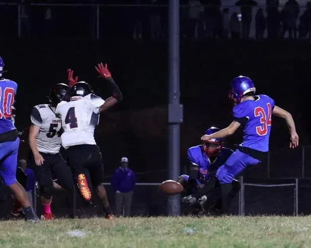 A blue-uniformed player kicks a football during a night game while players in white jerseys watch from the sideline.