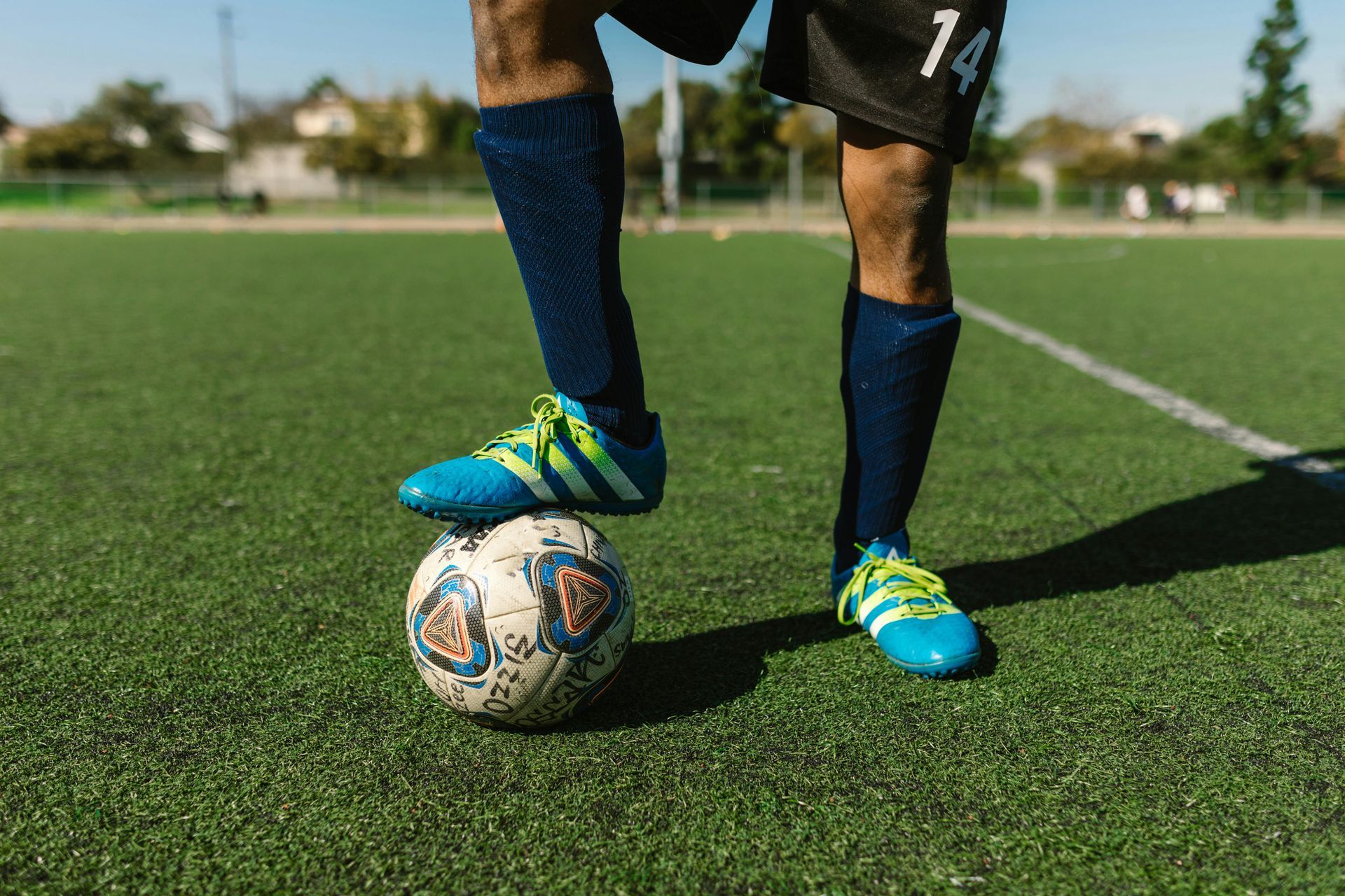A soccer player in black shorts and blue shin guards stands on a green field with one foot resting on a soccer ball.