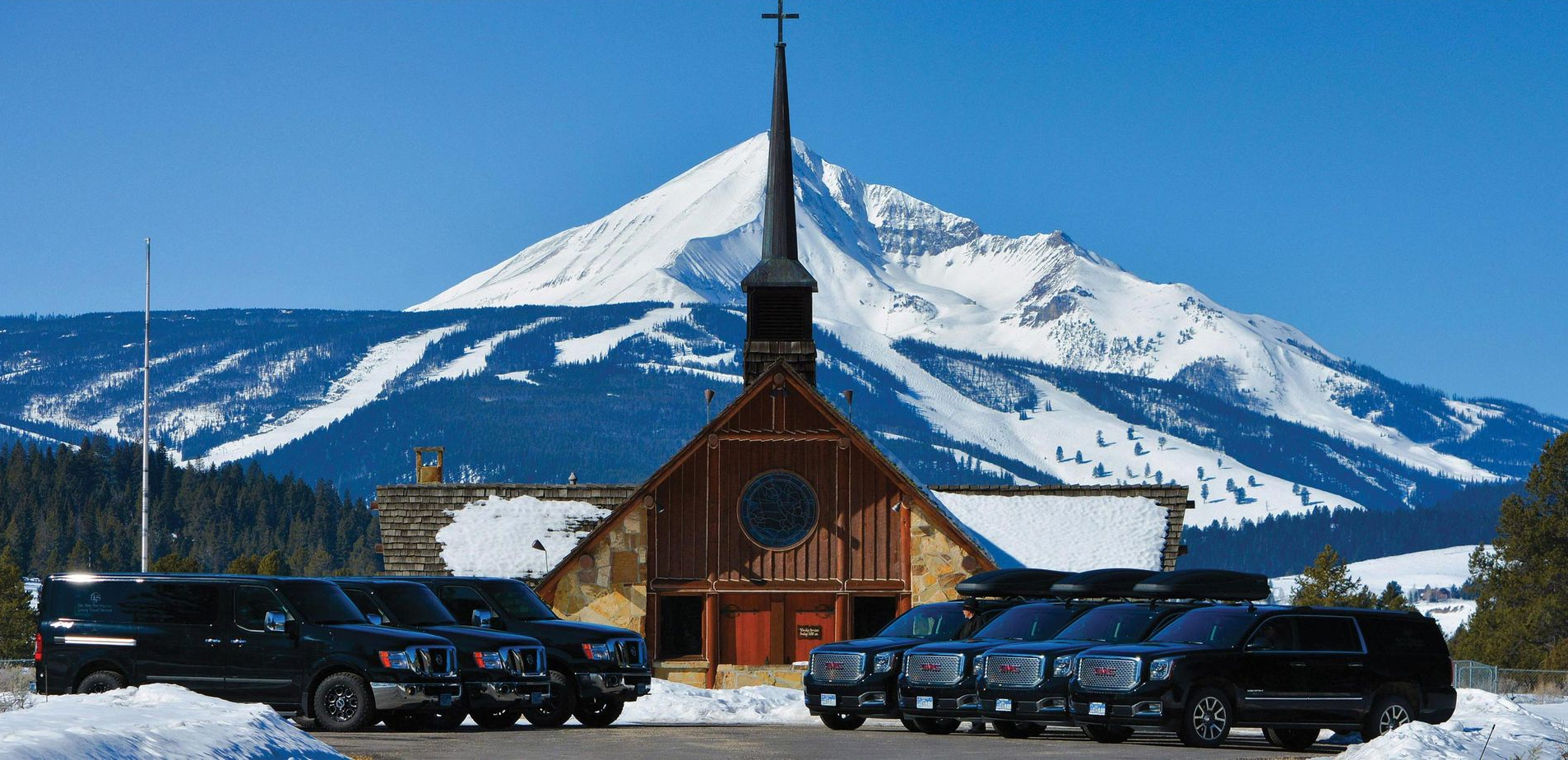Black vehicles parked in front of a church with a snowy mountain backdrop.