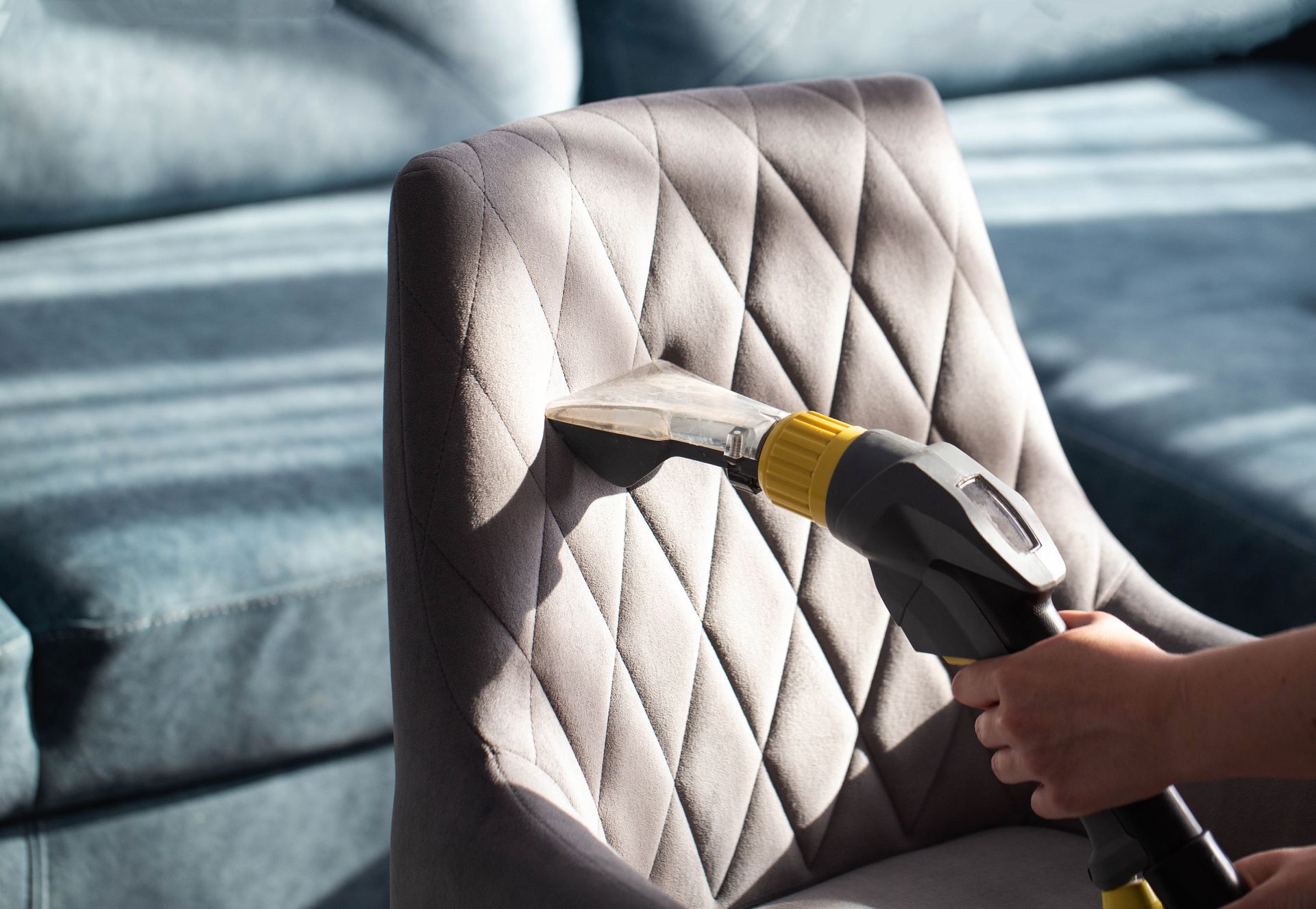Handheld vacuum cleaning a gray upholstered chair in sunlight