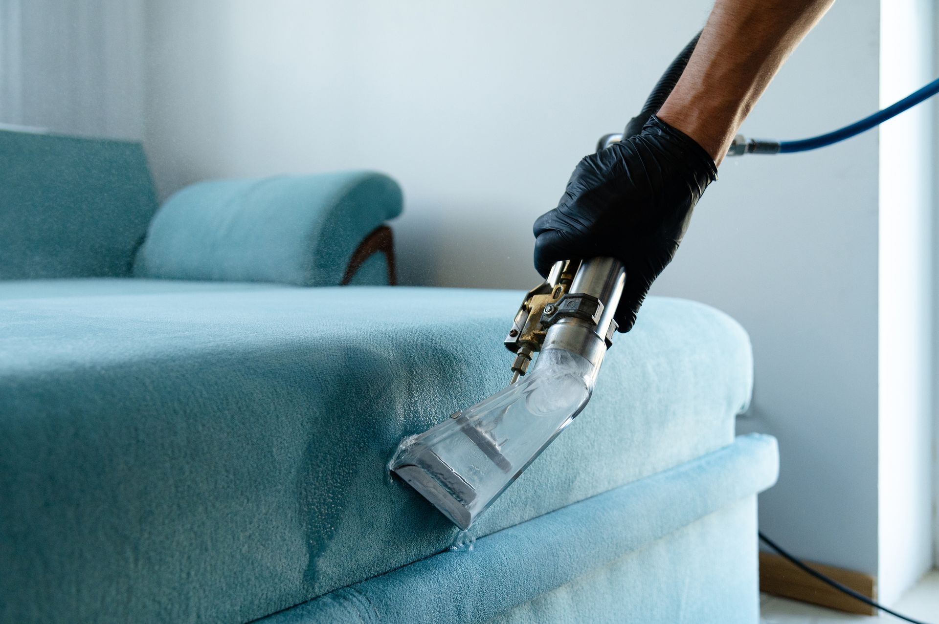 Handheld vacuum cleaning a light blue sofa in a bright room