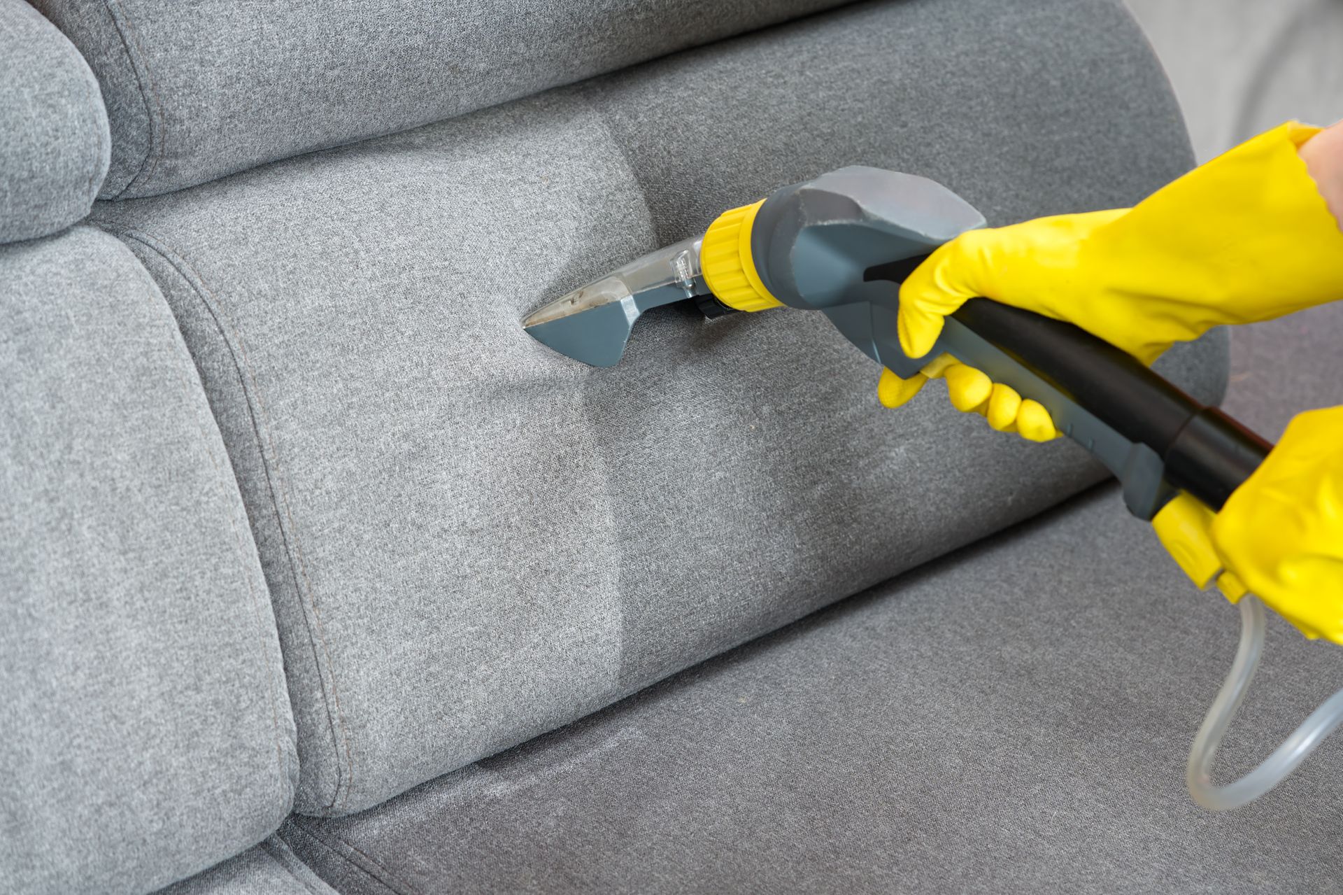 Yellow-gloved hand cleaning a gray sofa with a steam upholstery nozzle