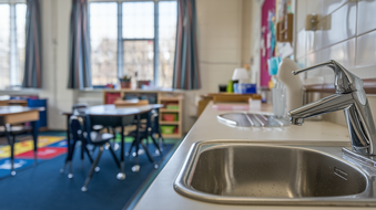 A kitchen sink in a classroom with tables and chairs in the background.