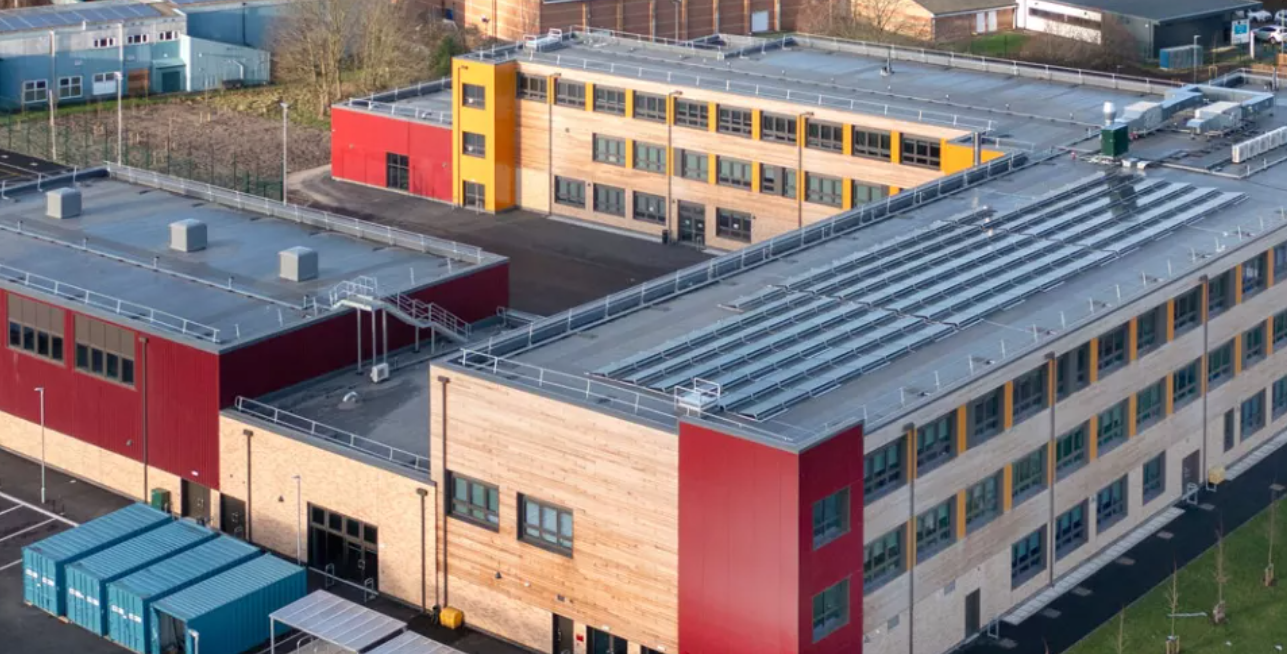 Aerial view of a large industrial building complex with red and beige walls and a rooftop lined with solar panels