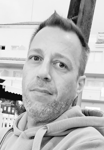 Man with short hair and stubble, looking at the camera. Wearing a hooded sweatshirt, outside.