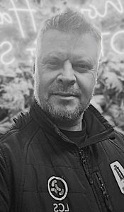 Man with beard, black jacket, smiling. Neon sign and foliage visible in the background.