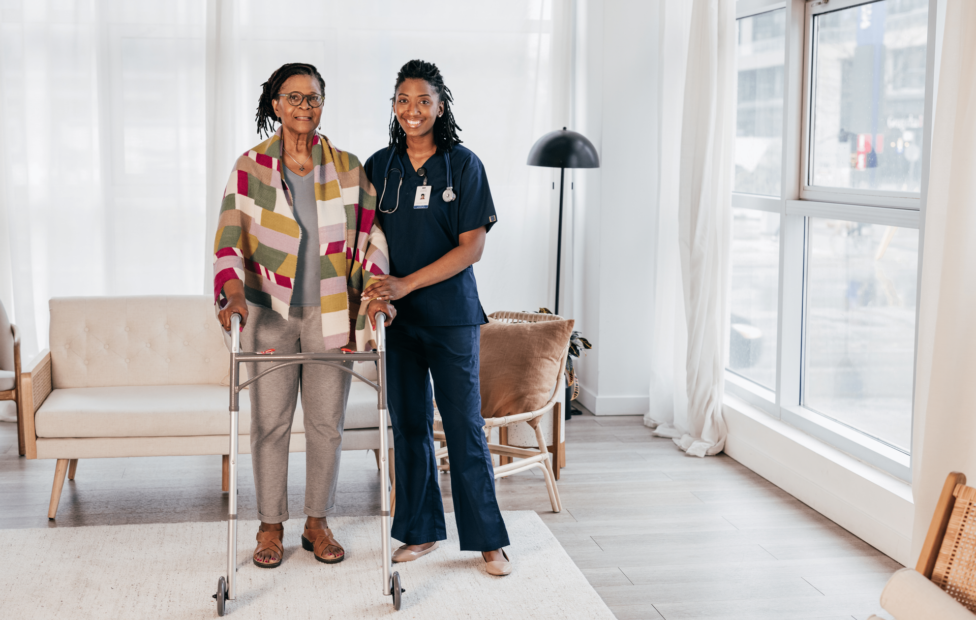 Een oudere zwarte vrouw met een rollator en een lachende zwarte verpleegster in een lichte kamer.