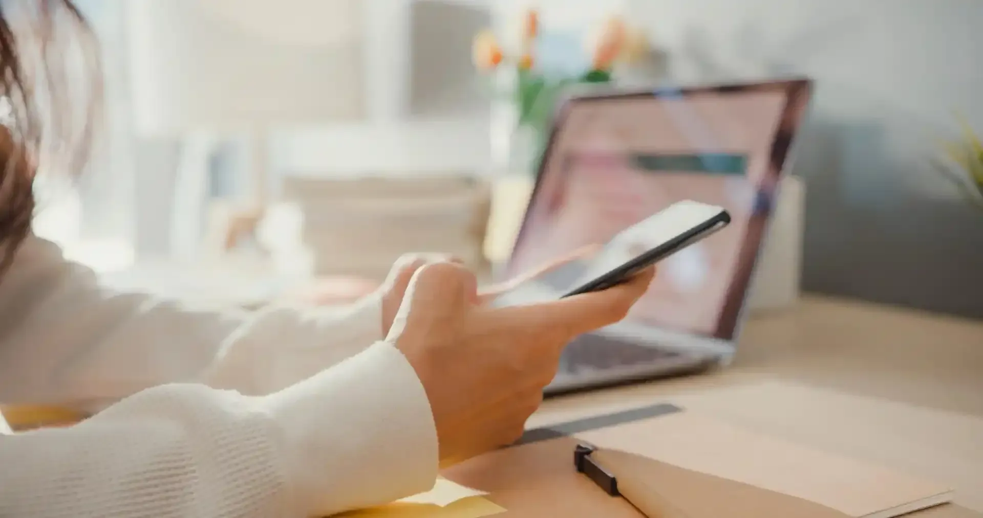 Persona che utilizza uno smartphone con un computer portatile sullo sfondo, in un ambiente chiuso.