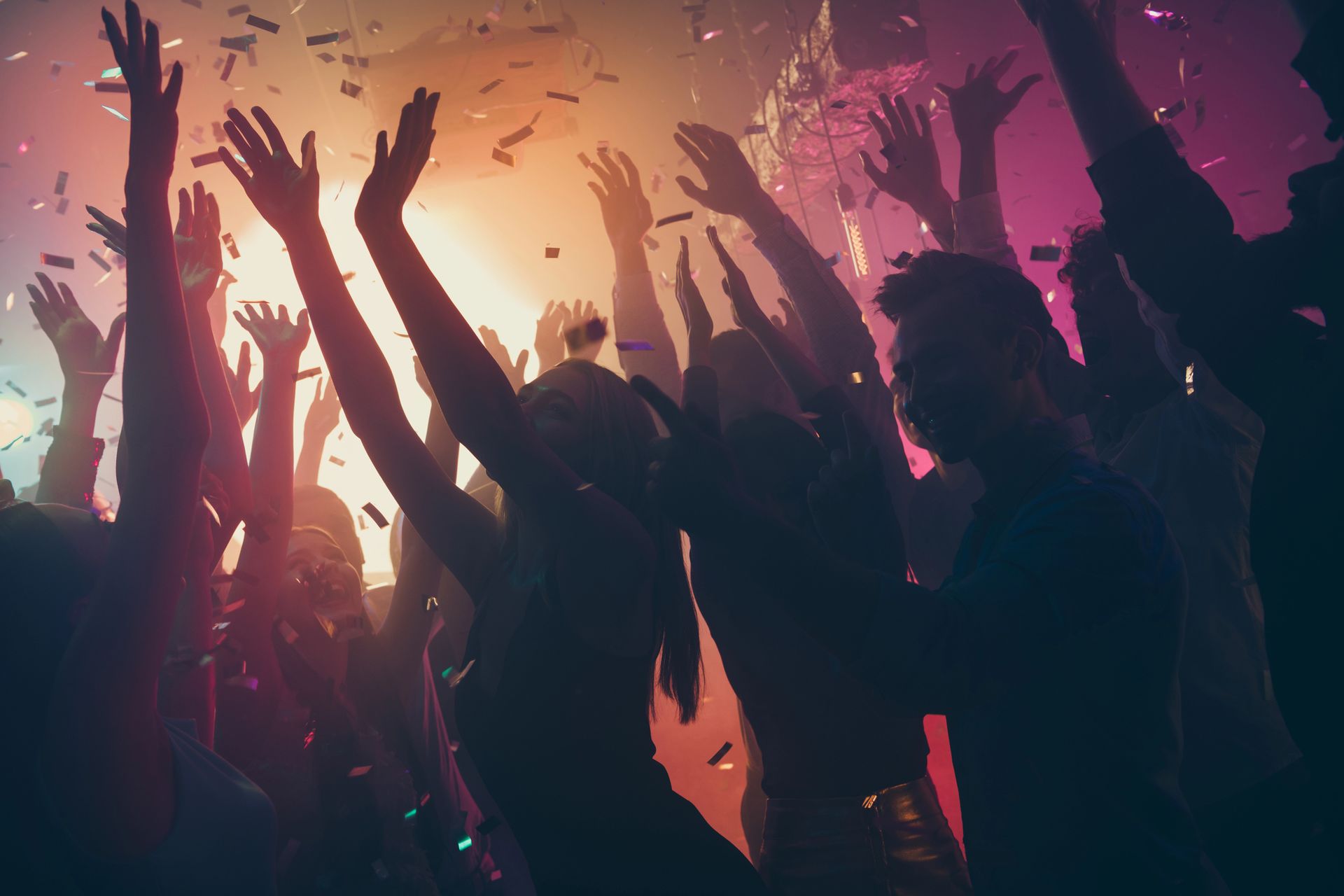 Persone che ballano a una festa con le mani alzate, coriandoli che cadono, luci colorate.