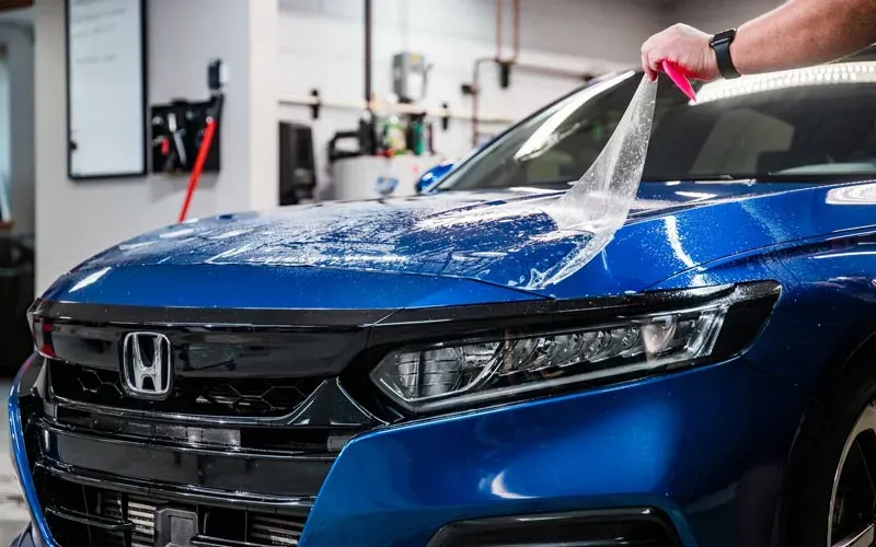 A person is applying a protective film to a blue Honda in a garage.