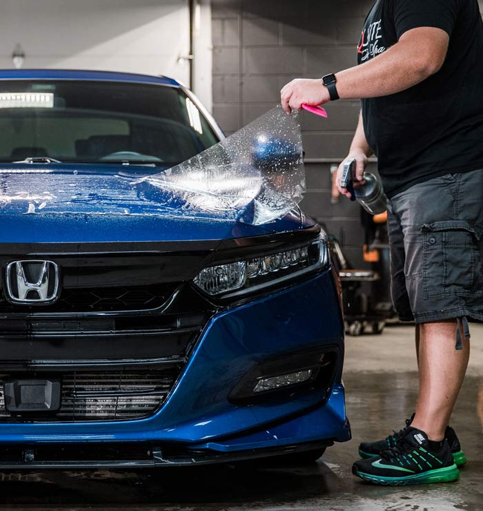 A person is applying a protective film to a blue Honda in a garage.