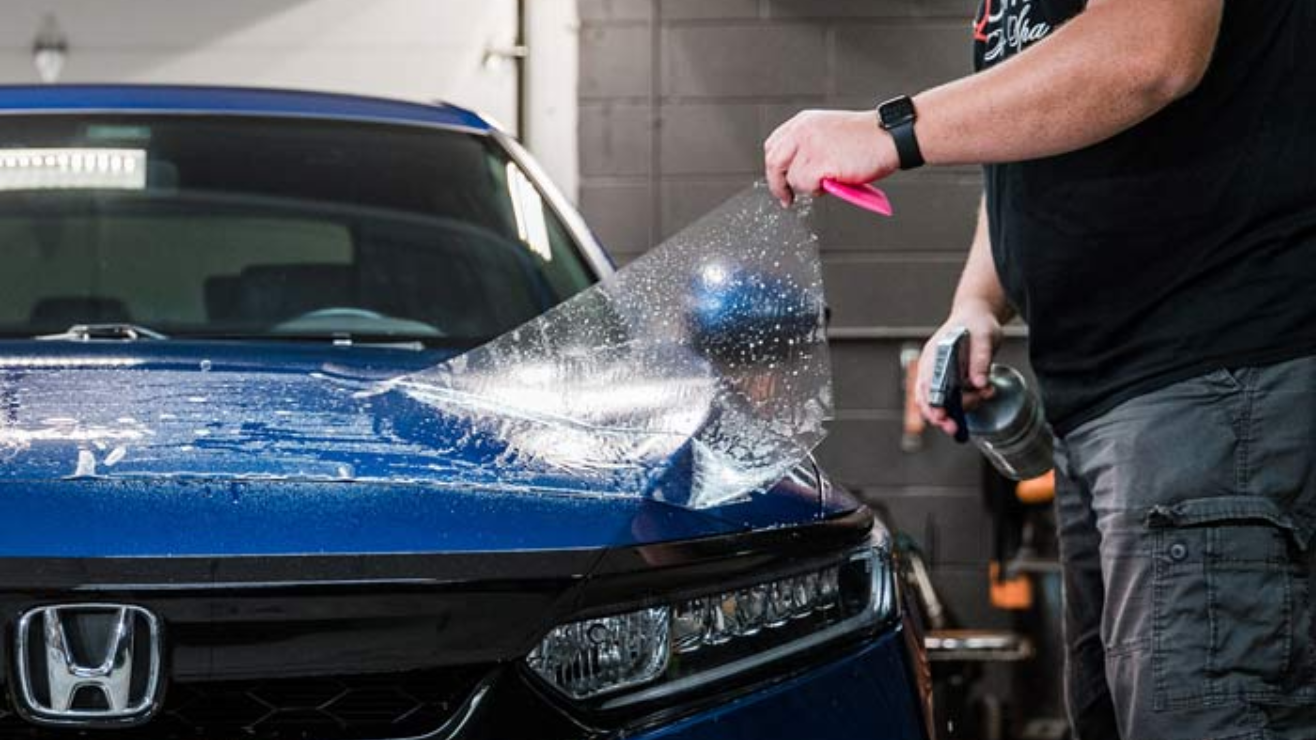 A person is applying a protective film to a blue Honda in a garage.