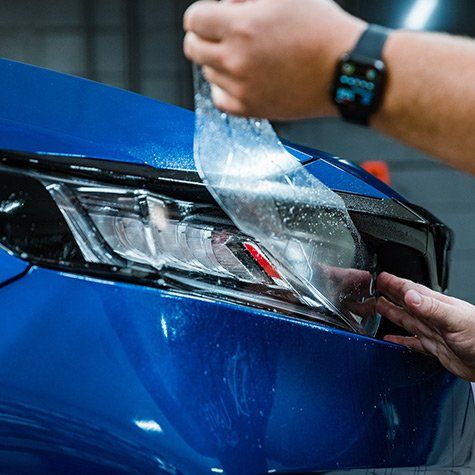 A person is applying a protective film to the headlight of a blue car.
