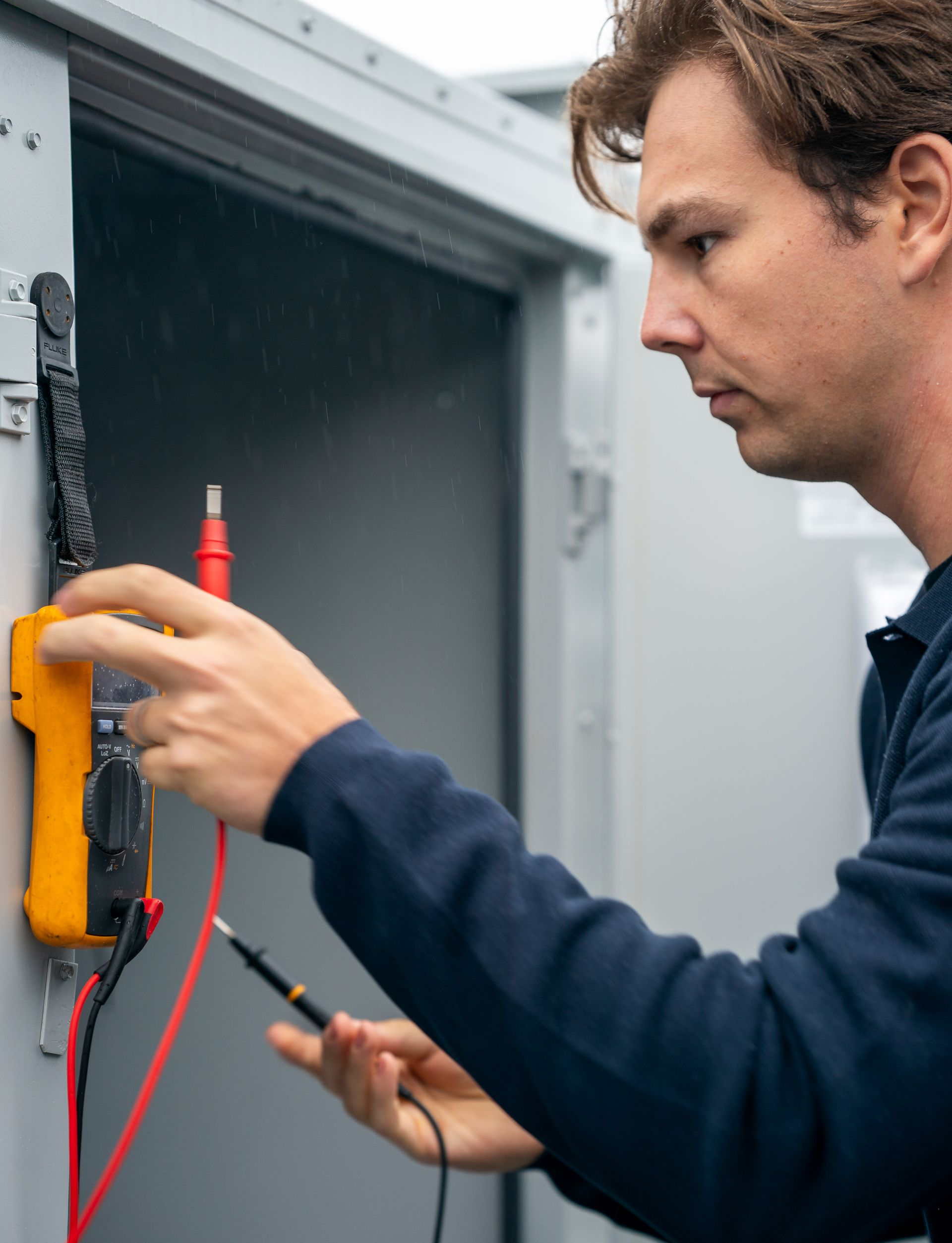 Man using a multimeter inside a metal cabinet, testing electrical components.