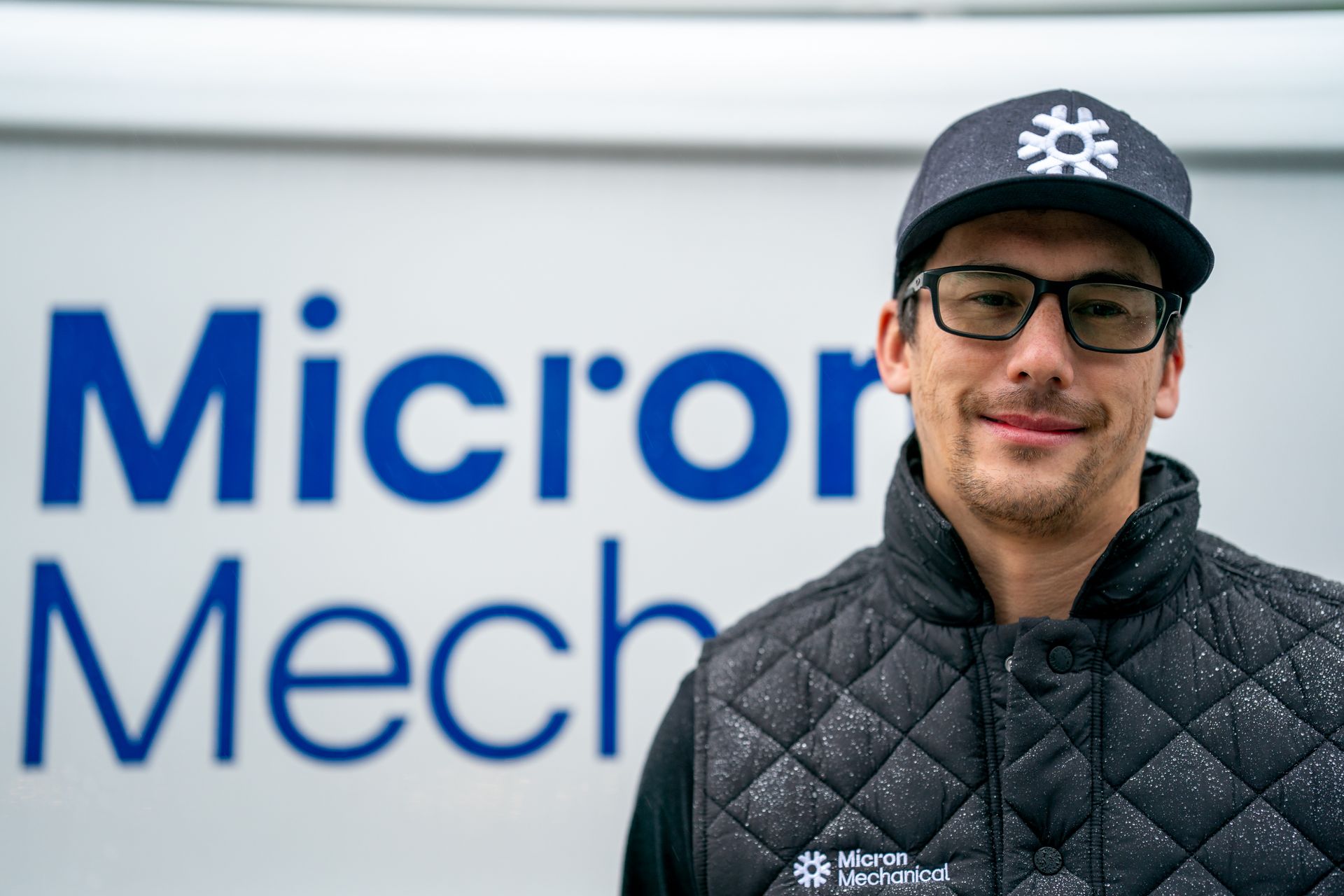Air conditioning unit next to a house with a wooden deck and stairs, surrounded by greenery.