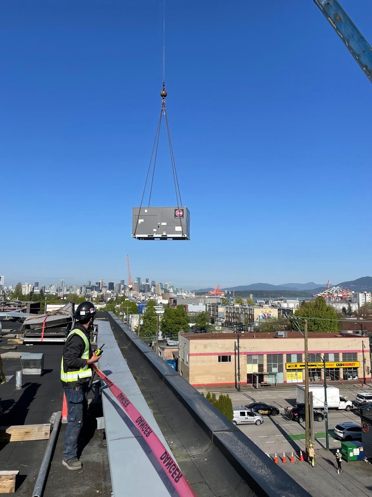 A worker on a rooftop watches a crane lift a large rectangular unit against a city skyline.