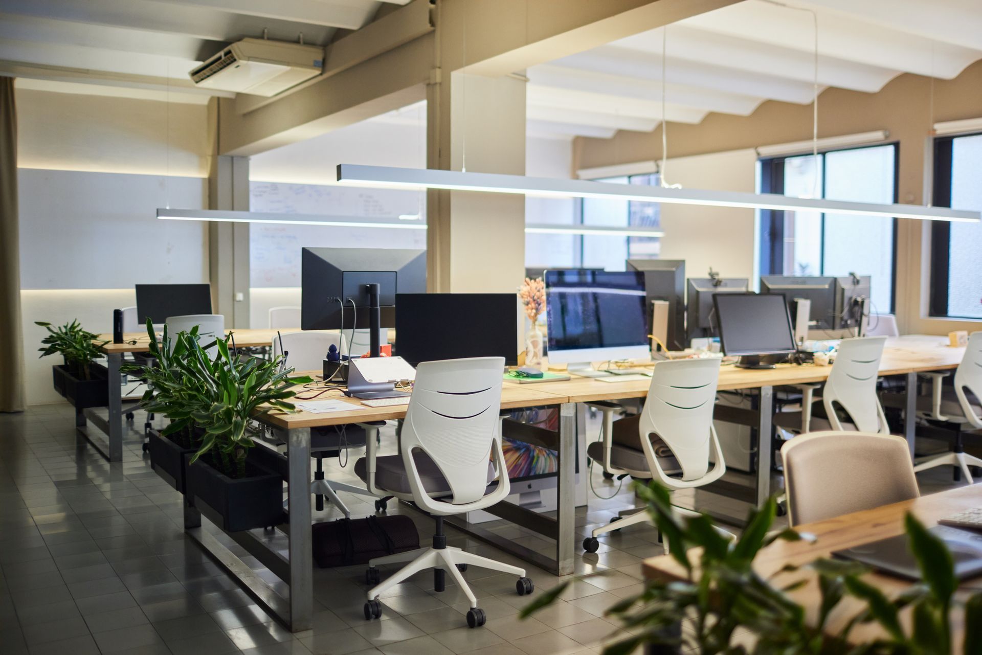 Modern office space with desks, computers, and white chairs. Indoor plants and overhead lighting.