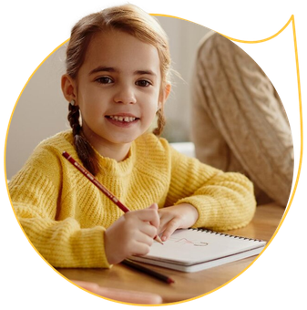 A little girl in a yellow sweater is sitting at a table writing in a notebook.