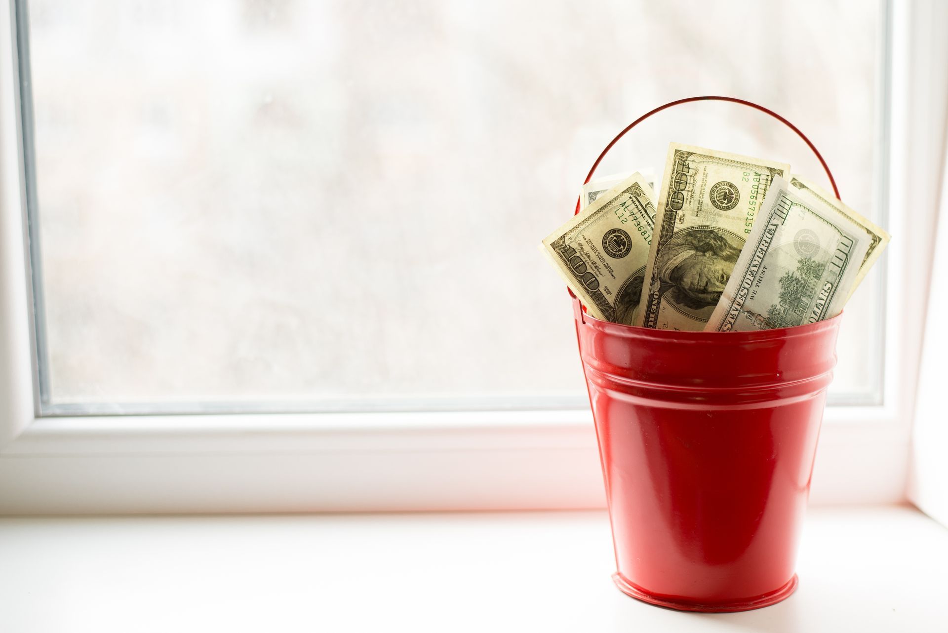 A red bucket filled with money is sitting on a window sill.