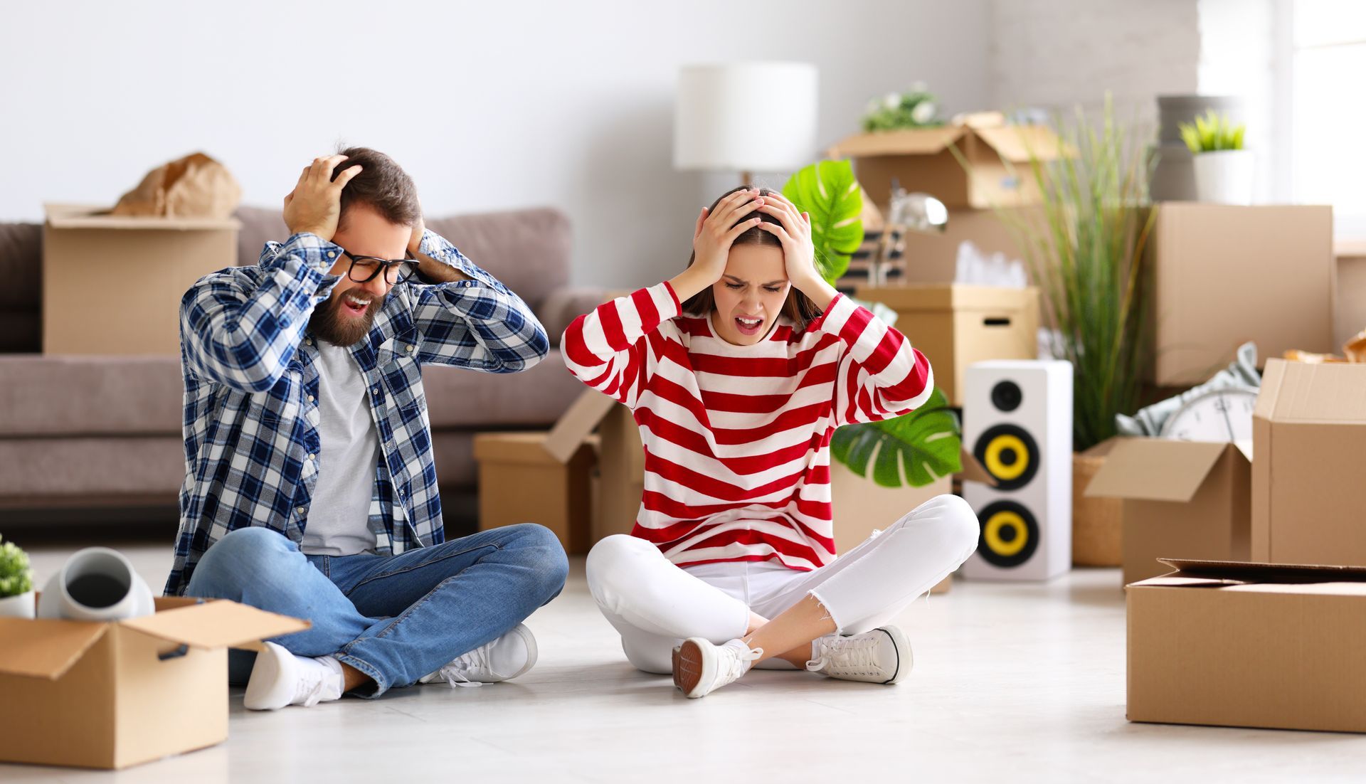 A man and a woman are sitting on the floor in a living room surrounded by cardboard boxes.