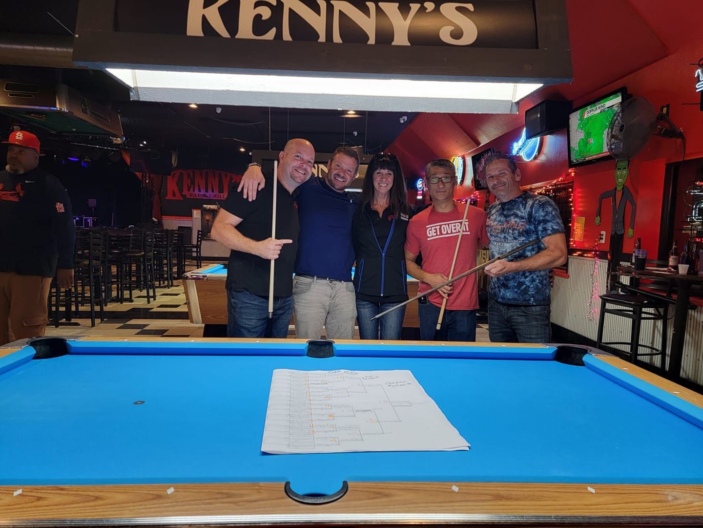 Five people stand together in a pool hall holding pool cues behind a blue billiard table with a paper sheet in the center.