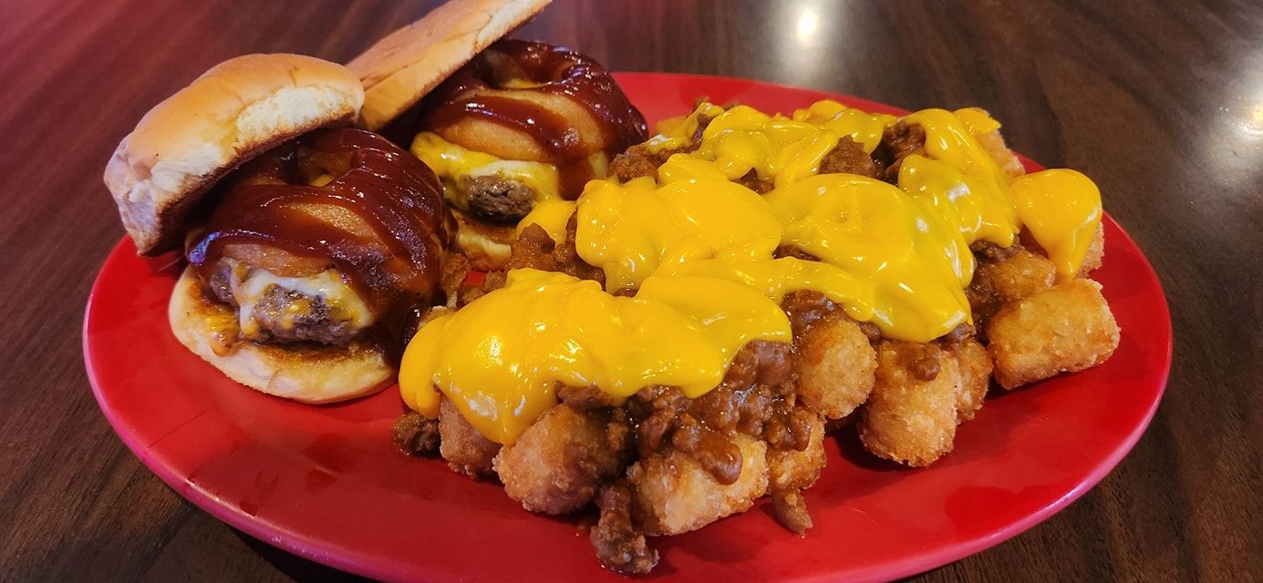 Two sliders topped with barbecue sauce beside a serving of tater tots smothered in chili and melted yellow cheese.