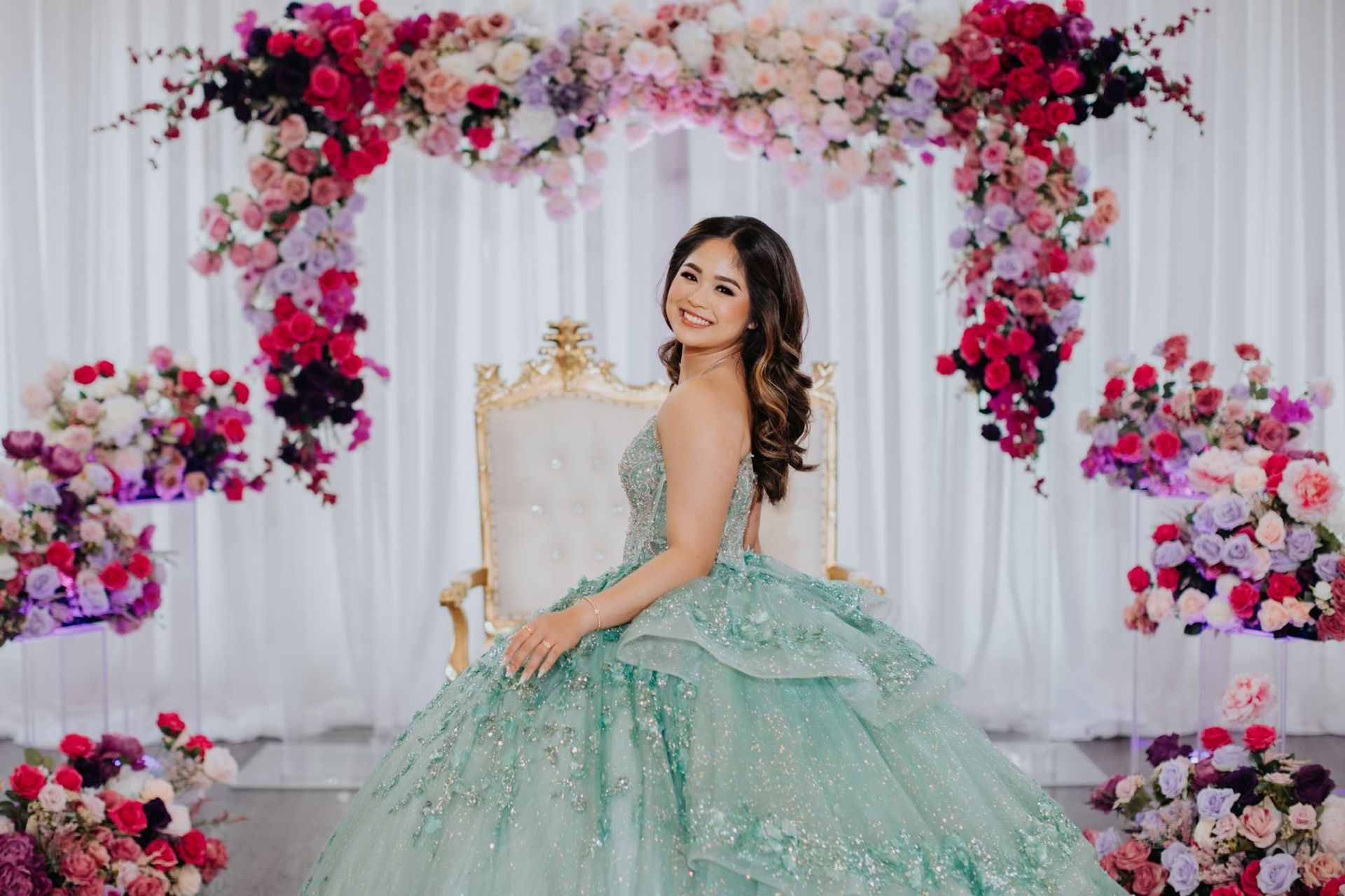 A woman in a green ball gown is sitting in a chair in front of a floral arch.
