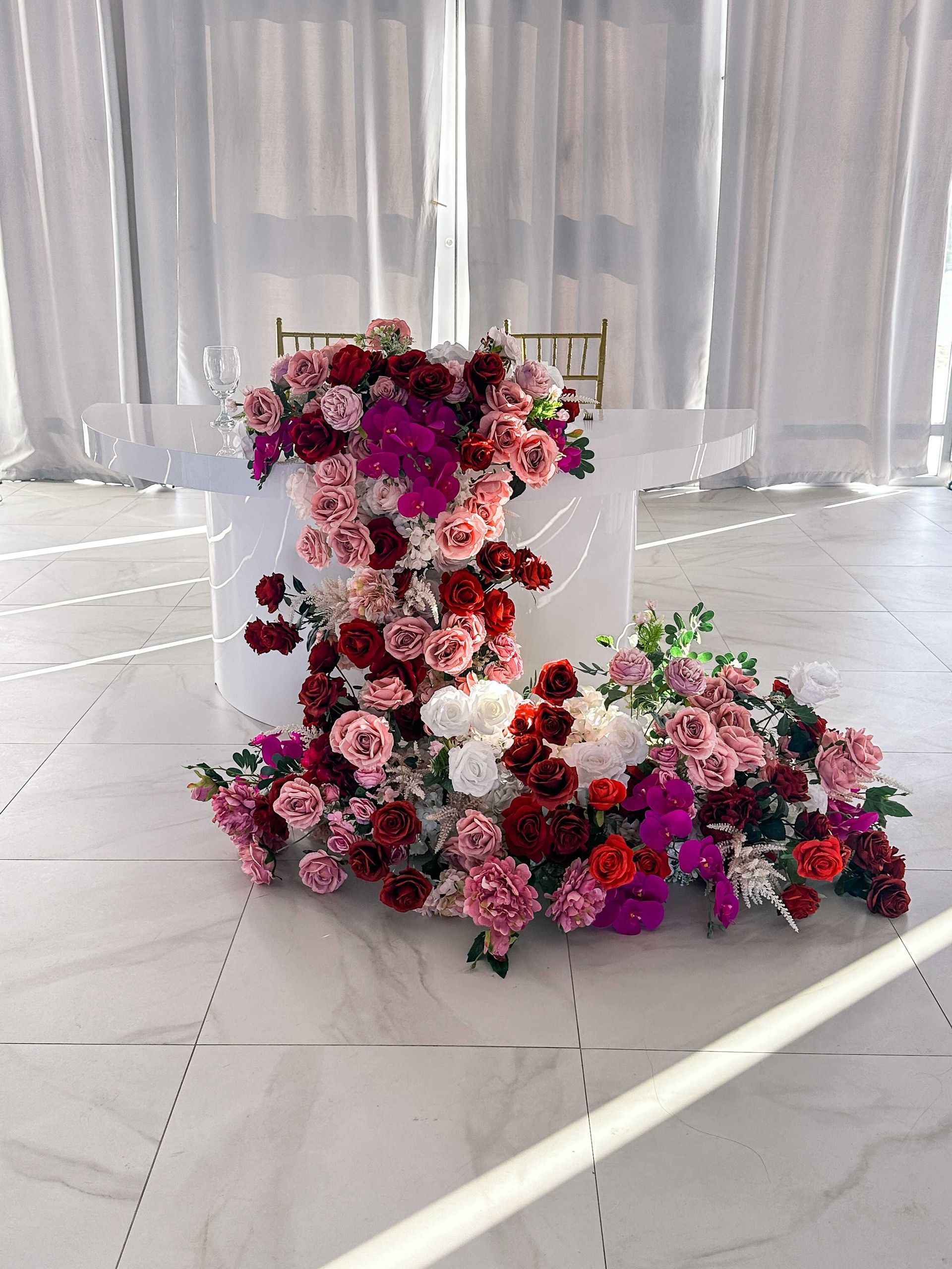 Wedding sweetheart table with cascading pink and red floral arrangement on white table and backdrop.