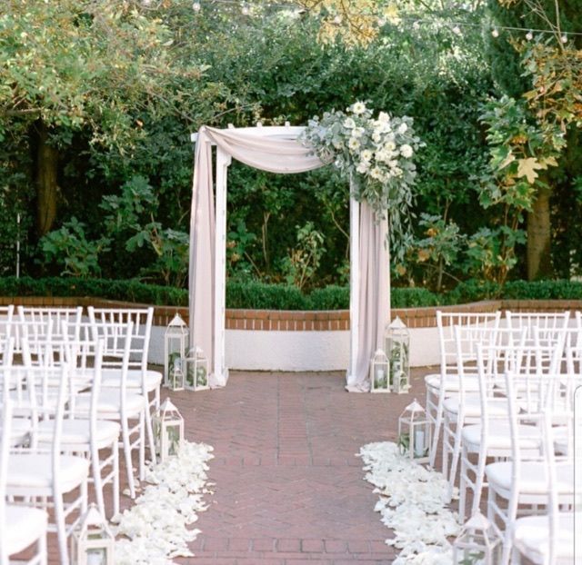 Wedding ceremony setup with white chairs, arch draped with fabric, and floral arrangement.