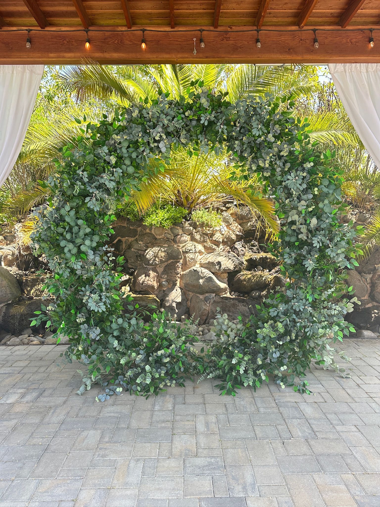 Circular greenery archway outdoors, against stone backdrop, on a brick patio, under a wooden structure.
