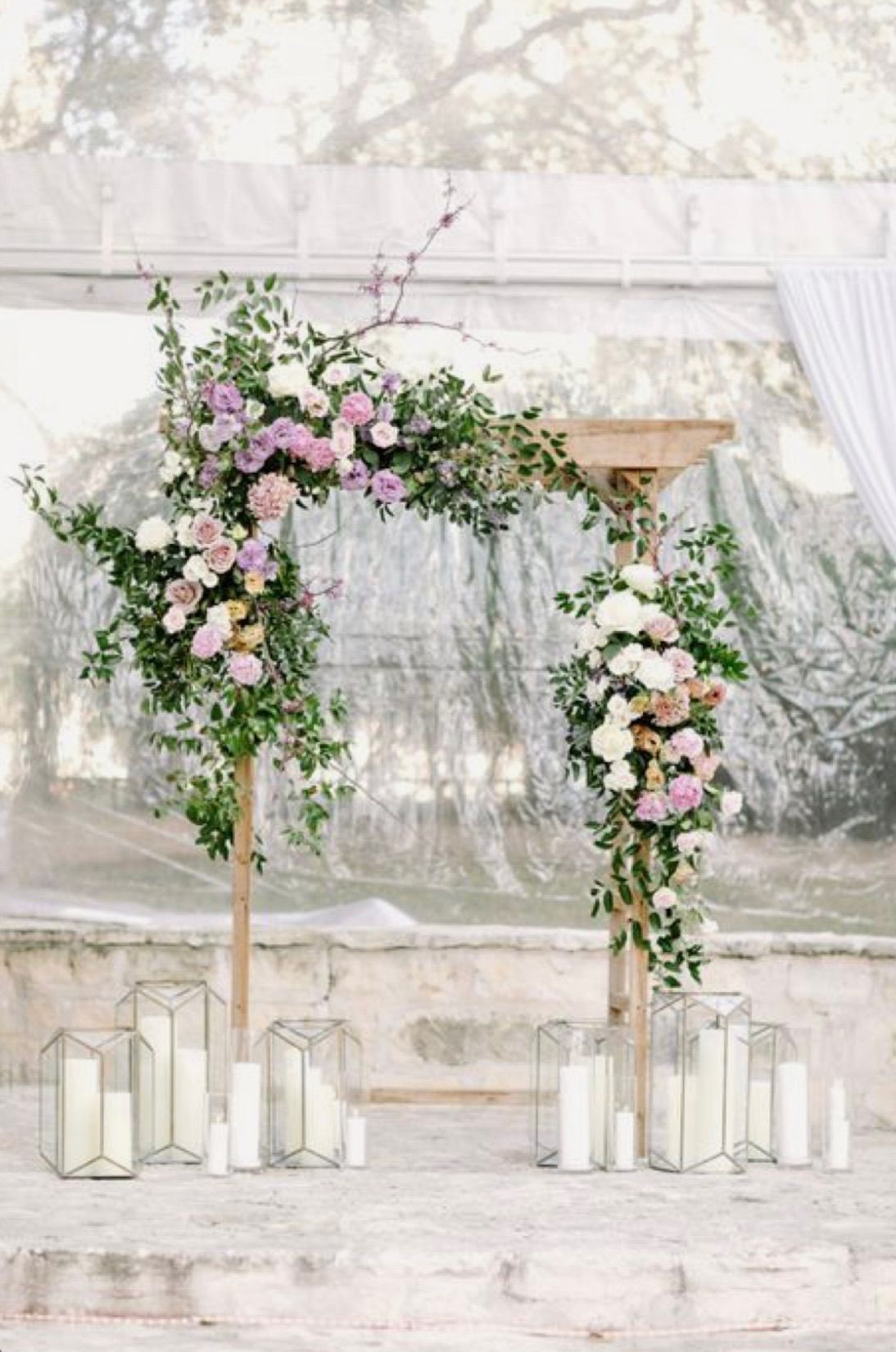 Wedding arch decorated with flowers and greenery, in front of a translucent backdrop with candles.