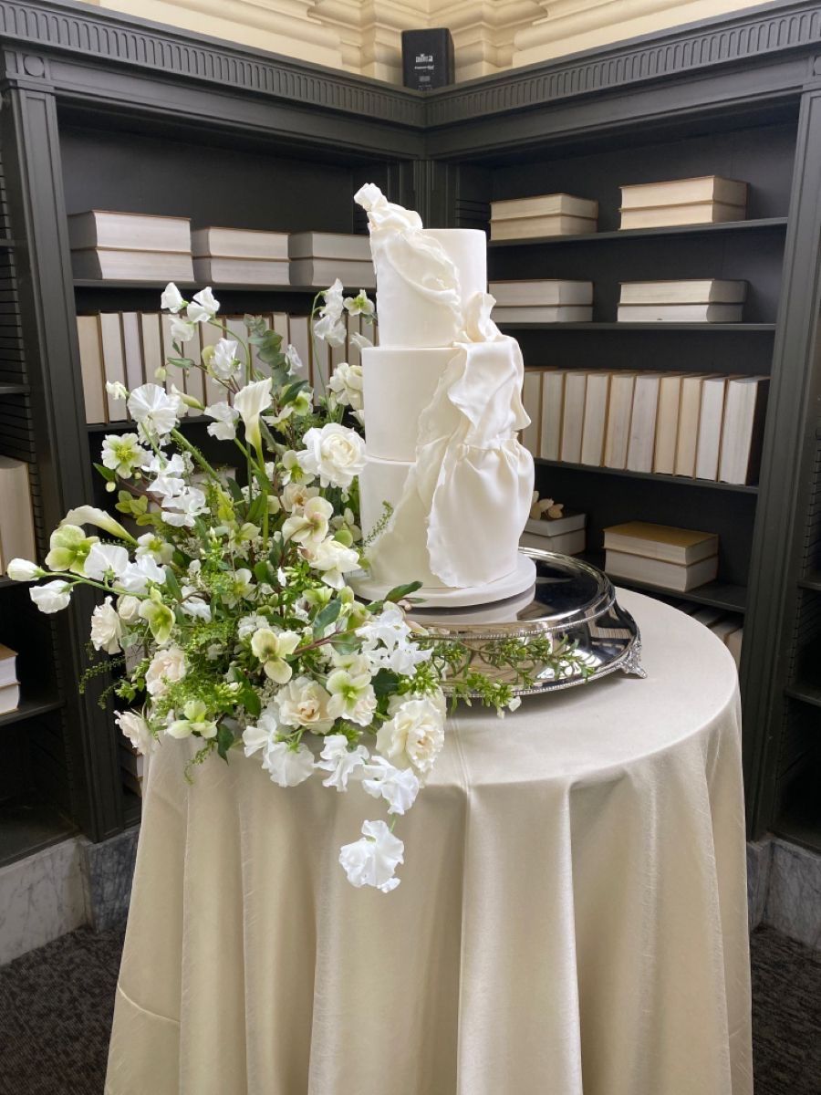 White tiered cake with floral arrangement on round table in library.