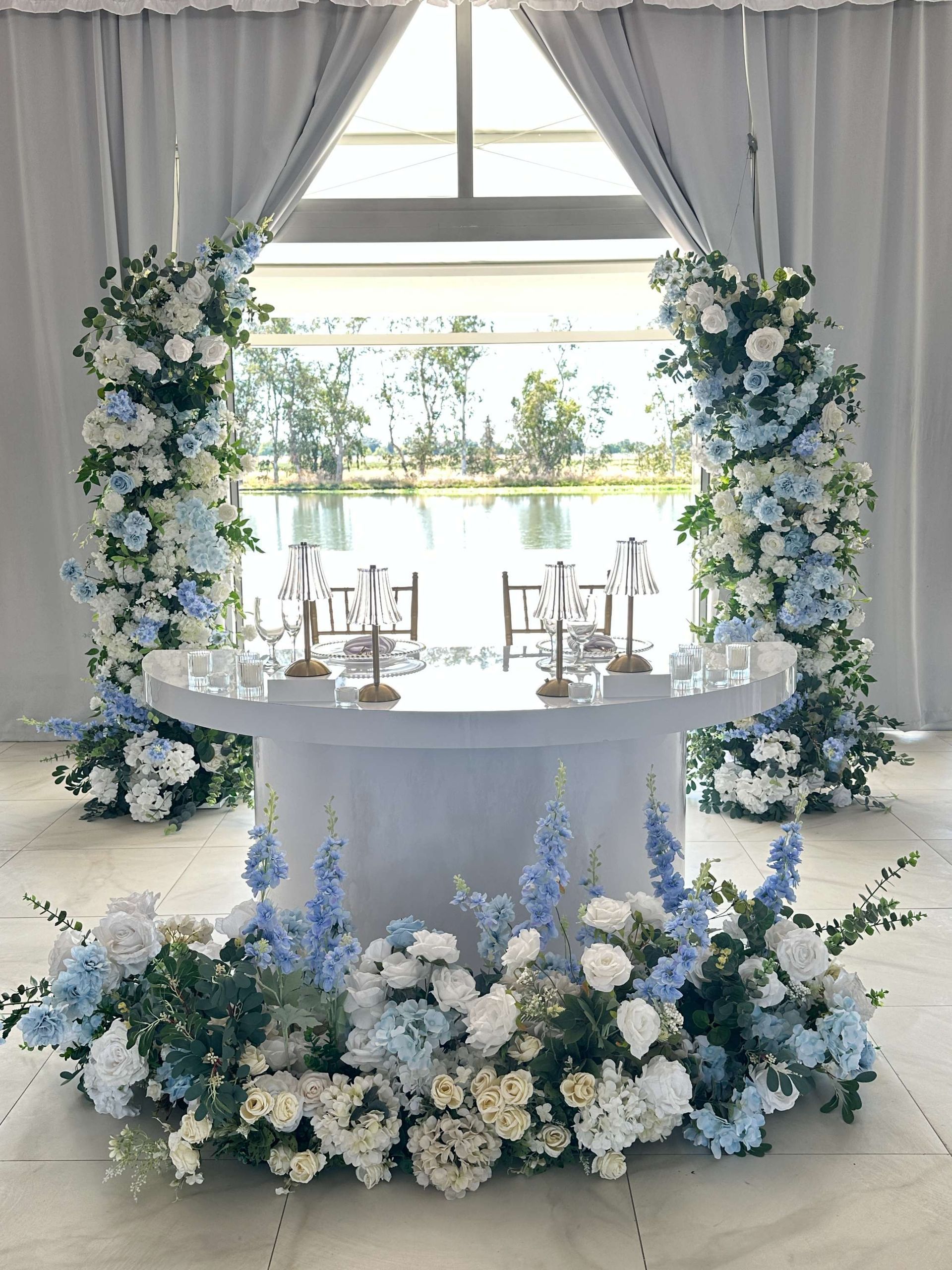 Floral-decorated wedding altar with white and blue flowers, two chairs, and a window overlooking water.