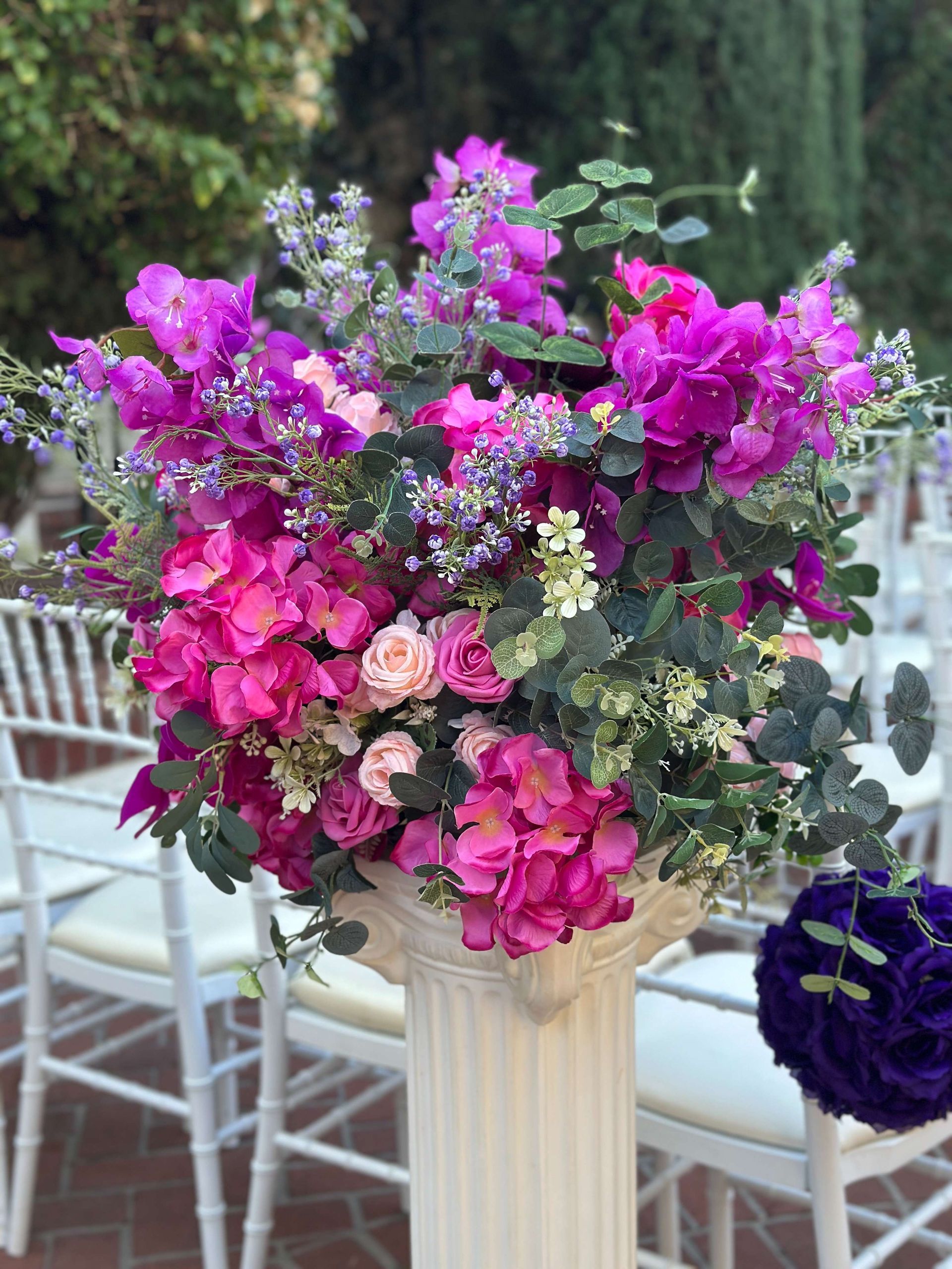Floral arrangement in shades of pink, purple, and green atop a white column, with white chairs in the background.
