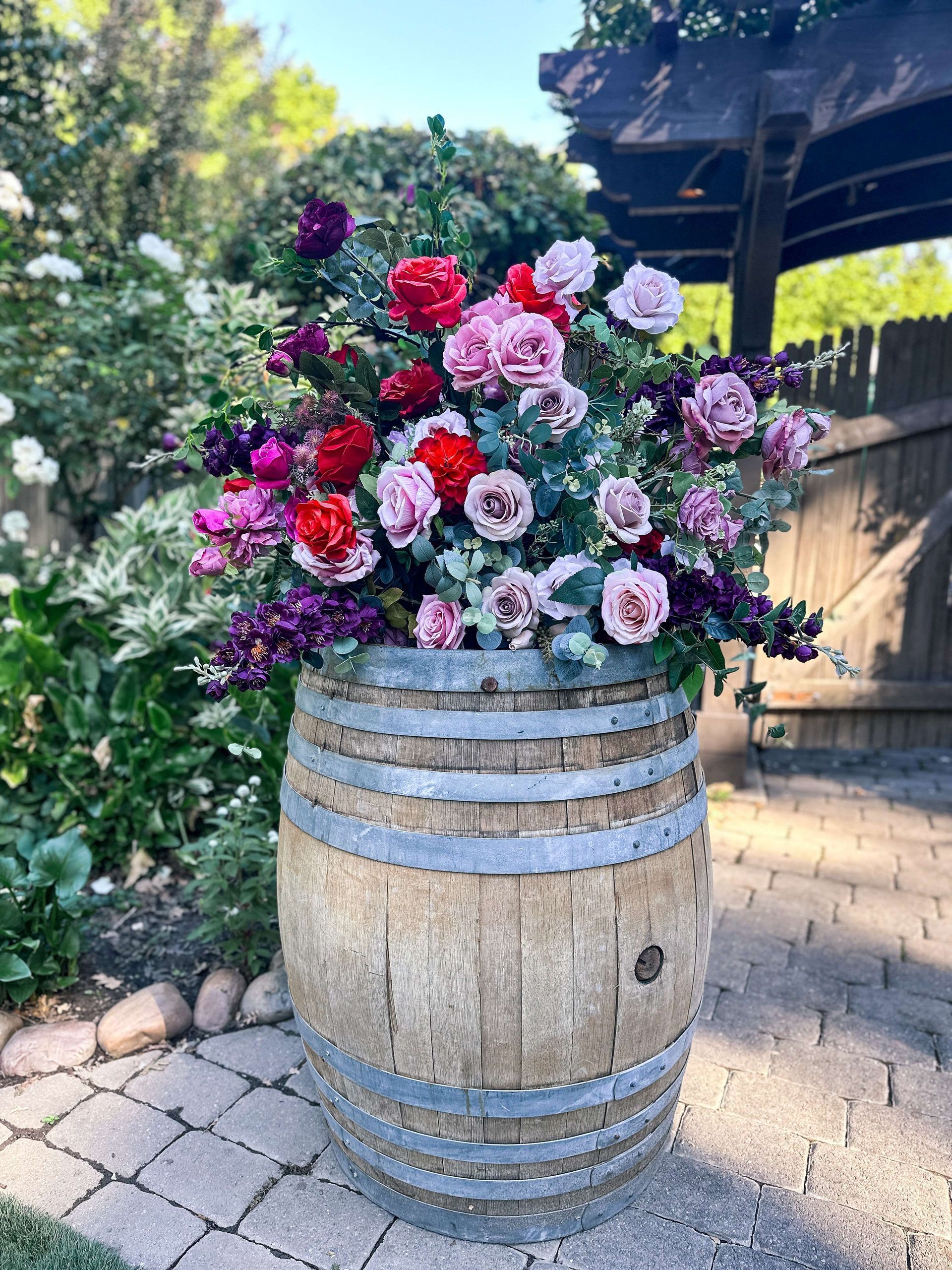 Flowers in shades of red, pink, and purple overflow a weathered wooden barrel outdoors.