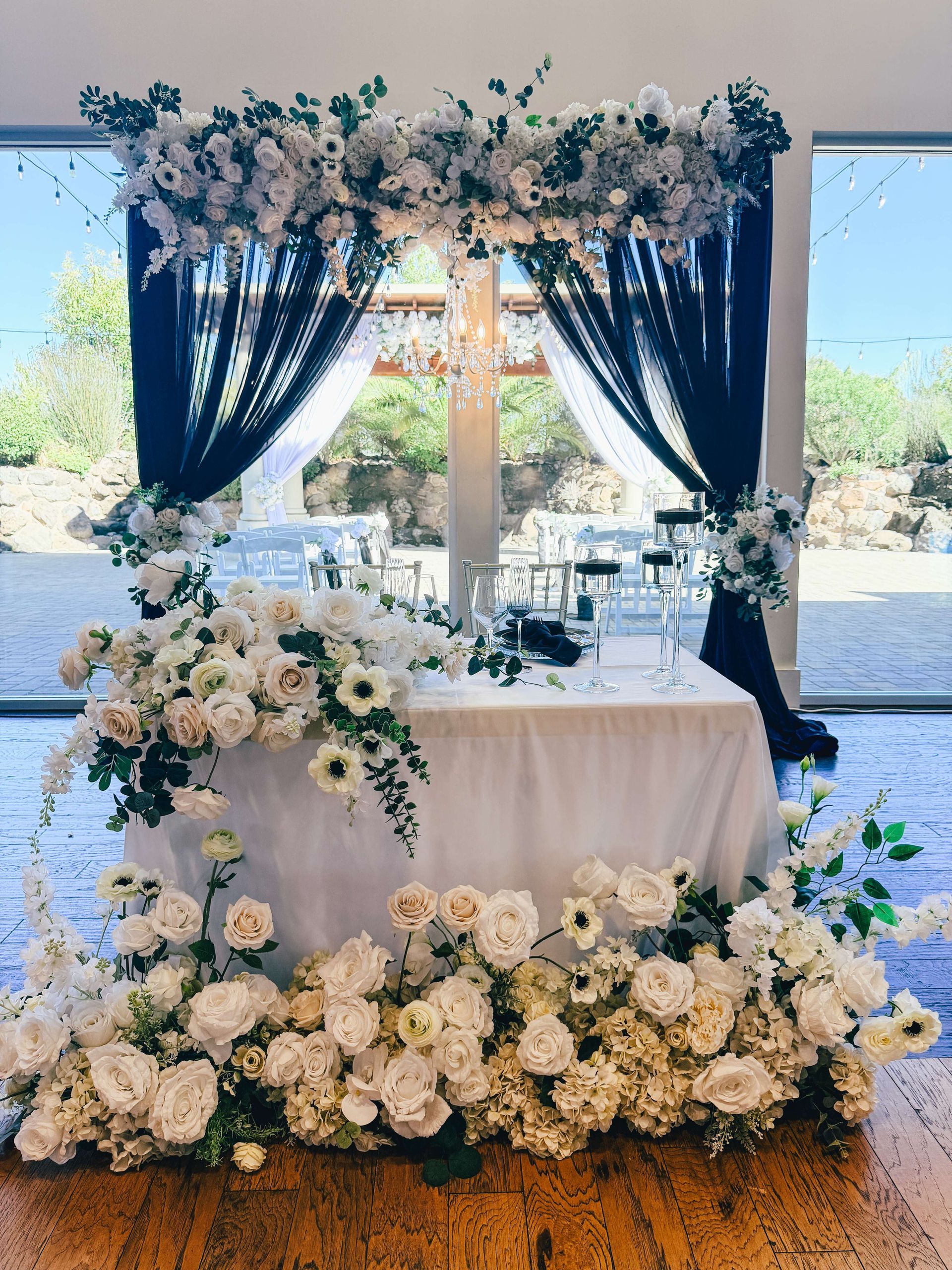 Wedding sweetheart table with white flowers, black drapery, and a floral arch in front of a window.