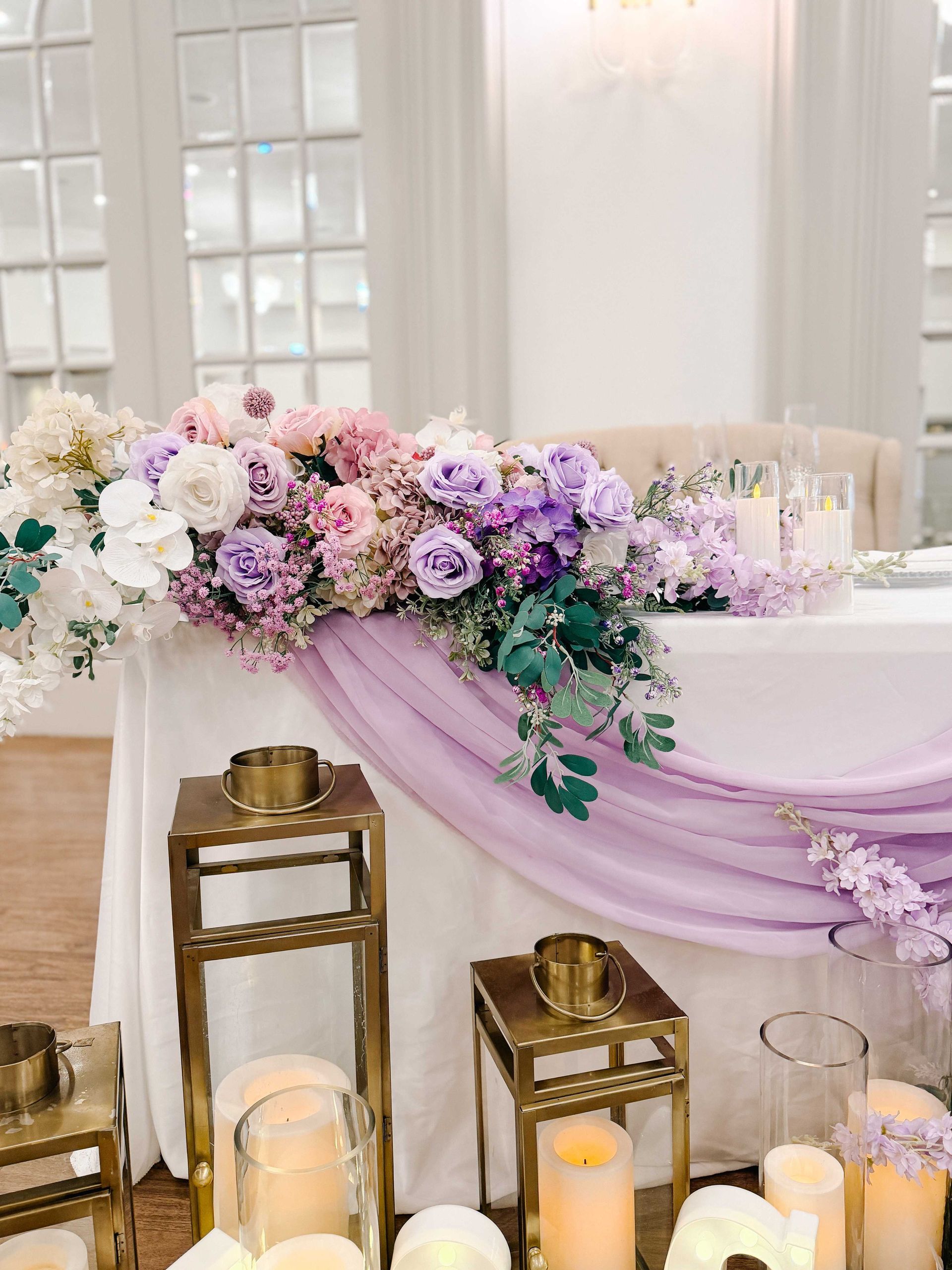 Wedding head table with lavender fabric, floral arrangement, gold lanterns, and candles.