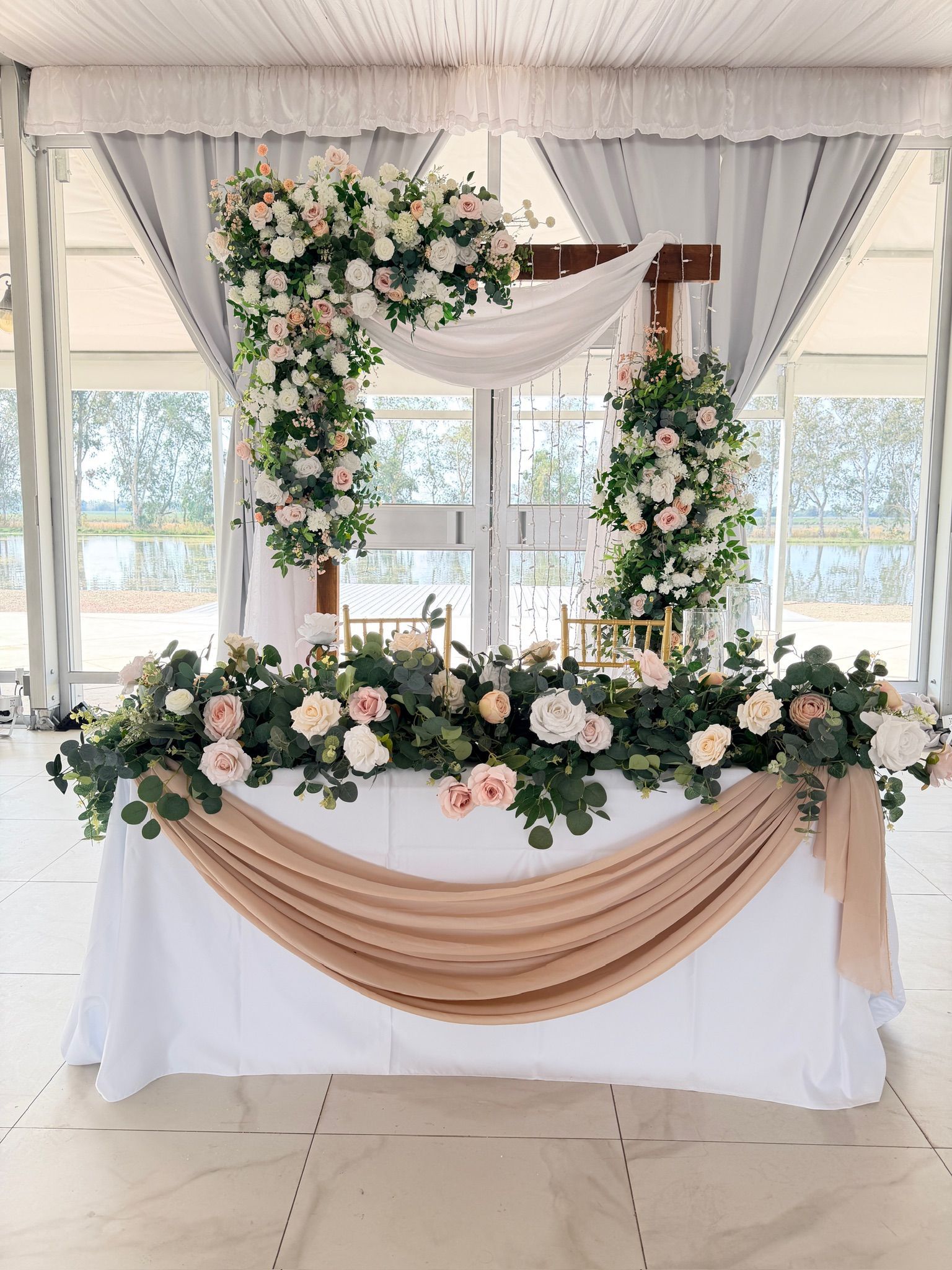 Wedding reception table decorated with flowers, draping, and floral arch in front of a window.