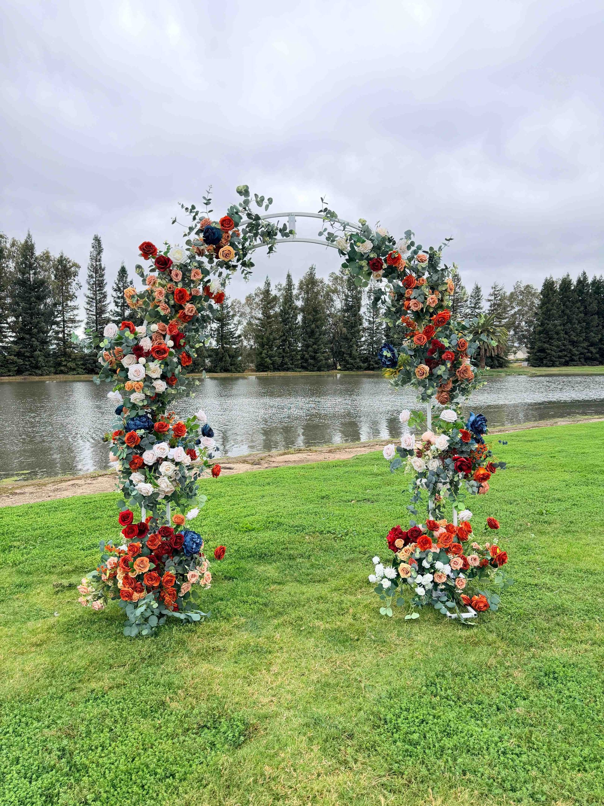 Floral arch with red, white, and blue flowers, on green grass, next to water and trees.