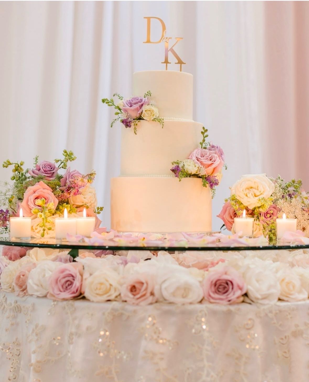 Three-tiered white wedding cake with floral accents and gold initials, surrounded by flowers and candles.