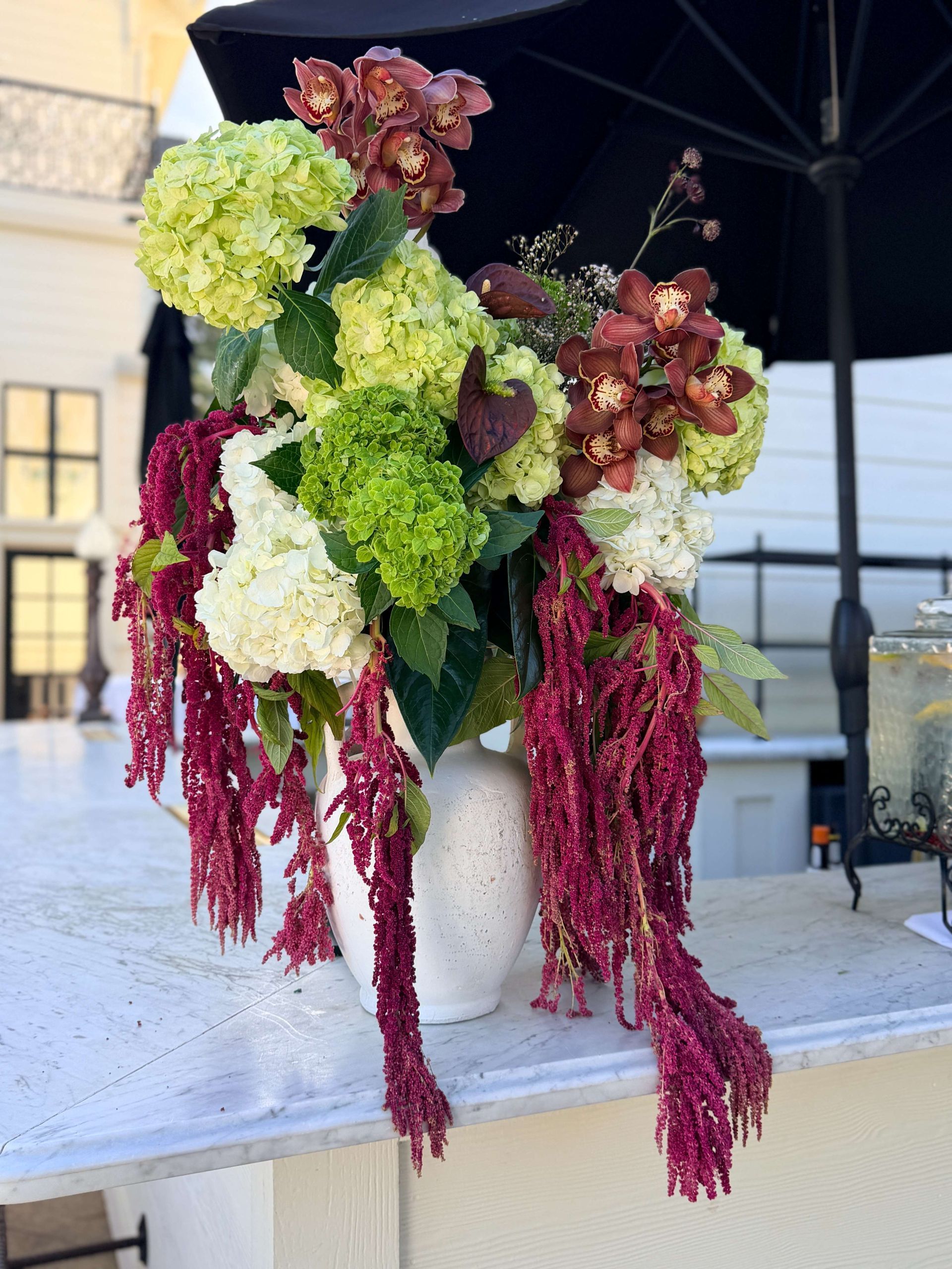 Floral arrangement in a white vase on a table with a black umbrella. Hydrangeas and amaranthus, with hints of red and green.