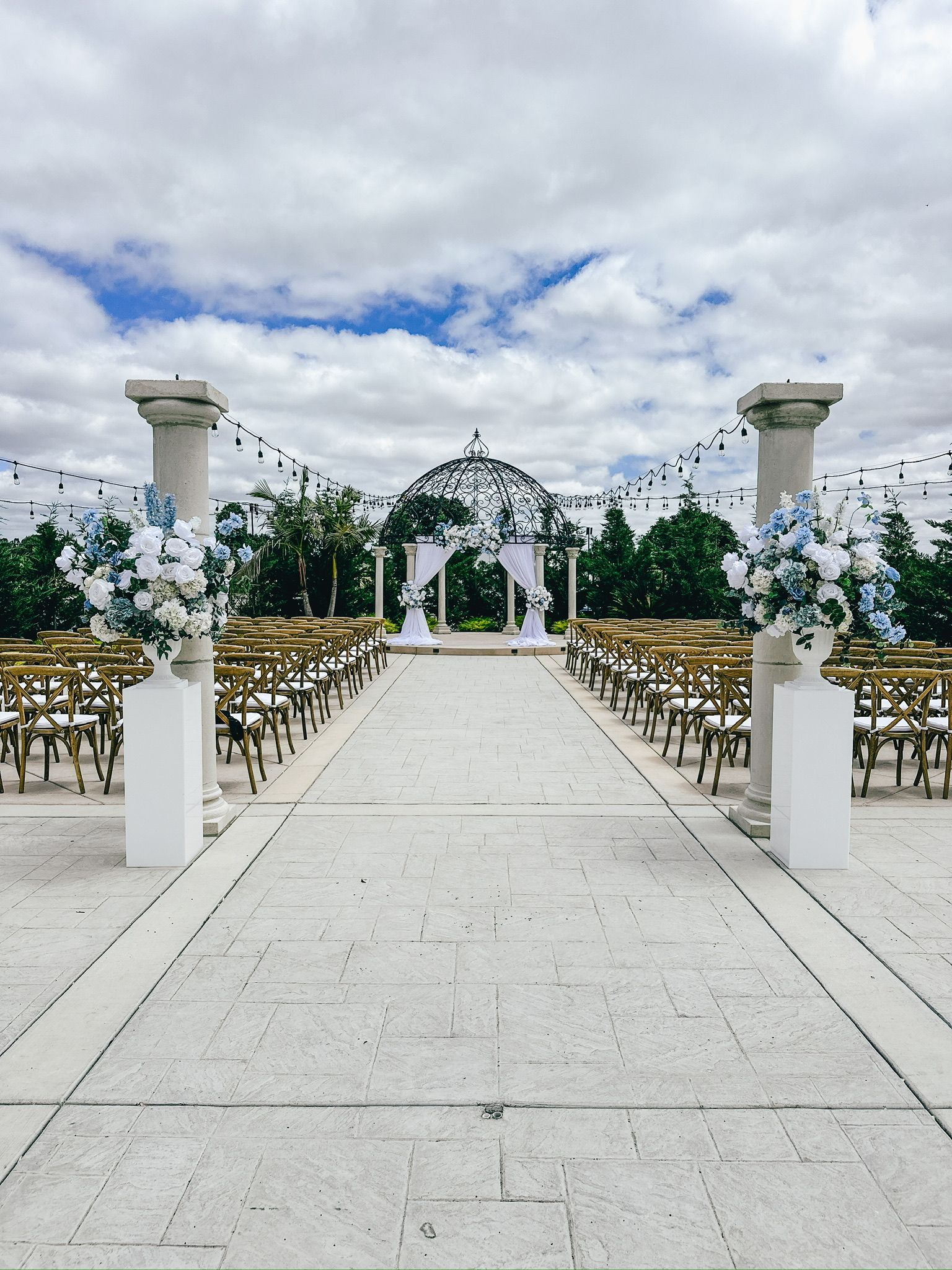 Wedding ceremony setup outdoors with blue and white floral arrangements, chairs, and archway under a cloudy sky.