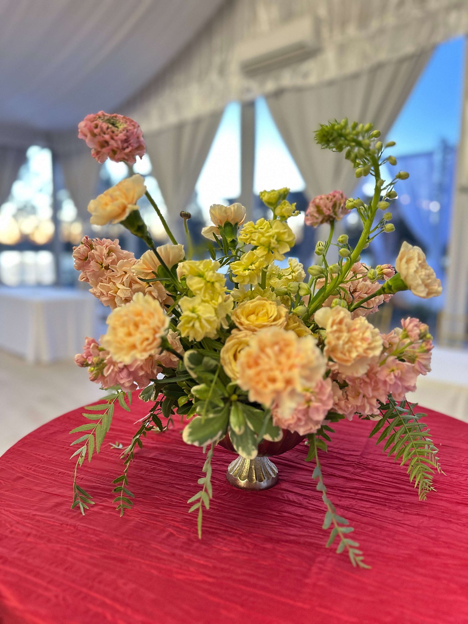 Floral arrangement on a red tablecloth; peach and yellow carnations, pink stock, and green accents in a silver vase.