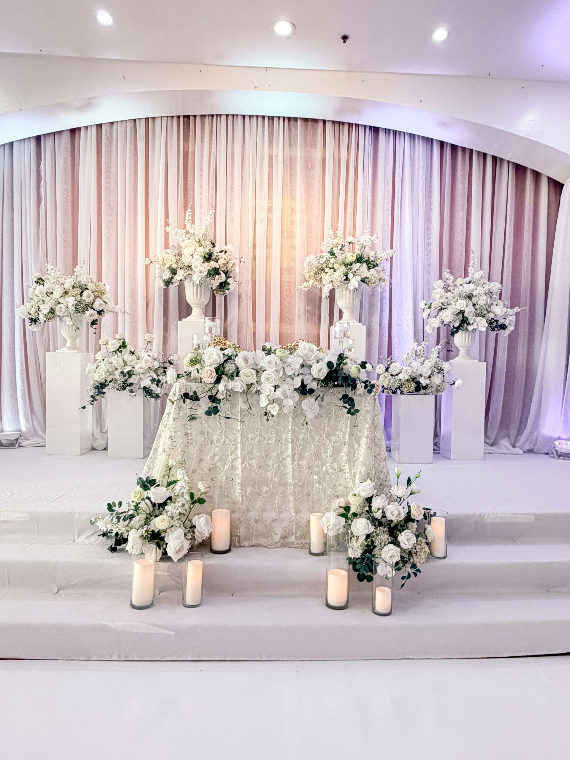 Wedding altar with white flowers on and around a decorated table and pedestals; light pink draped backdrop.