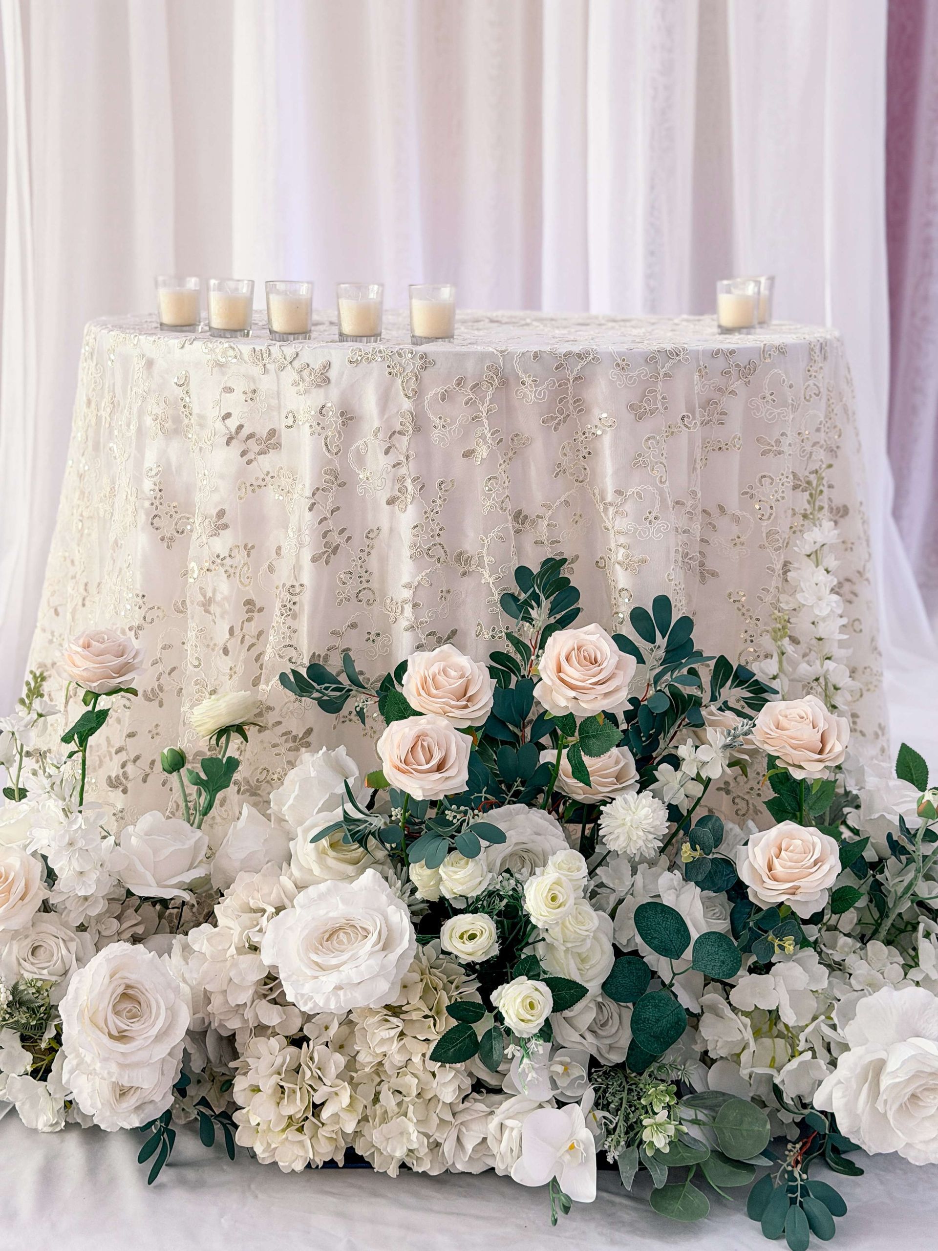 A round table with white floral arrangement and candles. Shimmering tablecloth.