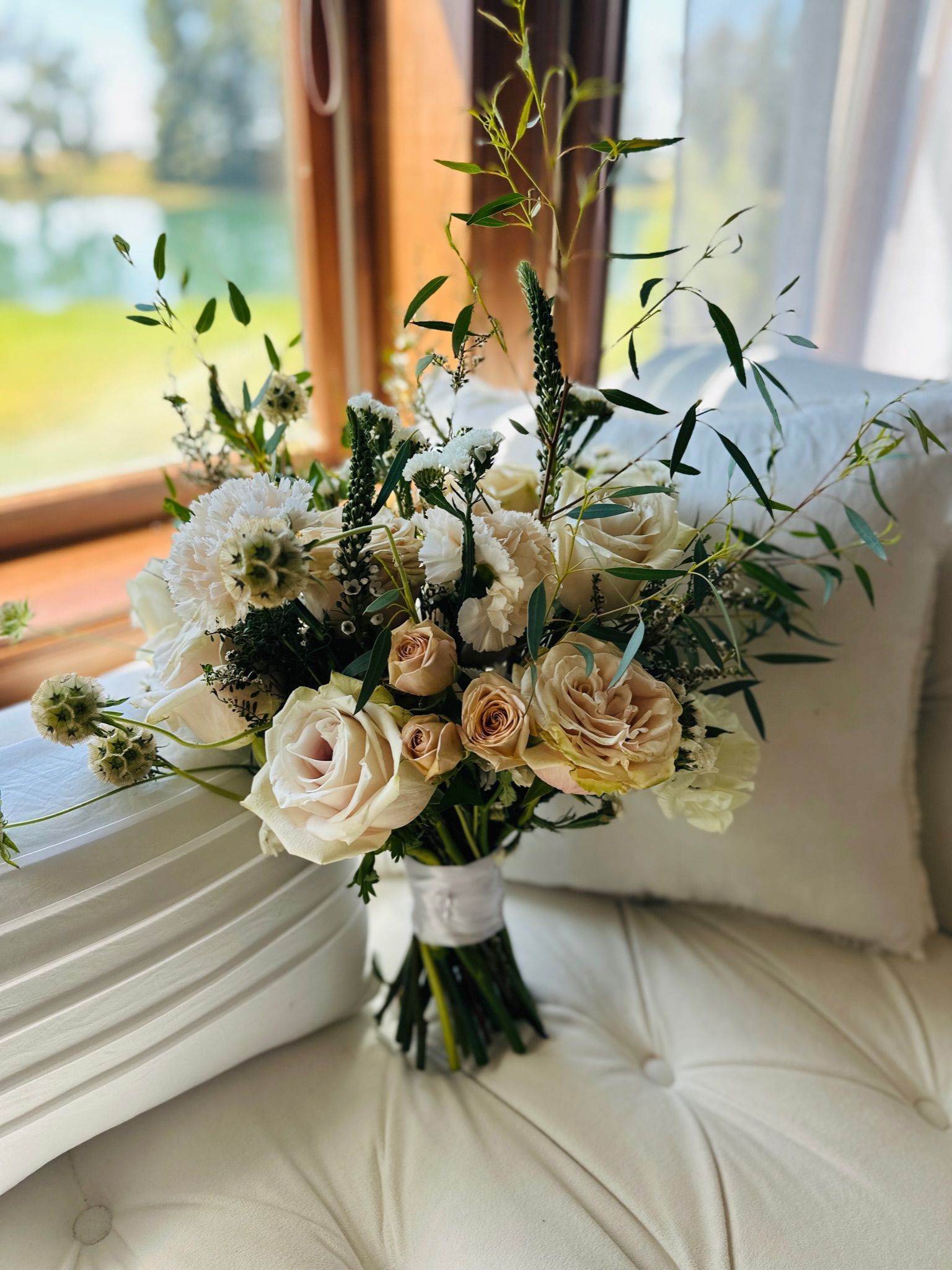 Bouquet of white and peach roses, hydrangeas, and greenery, tied with white ribbon, resting on a white cushion near a window.