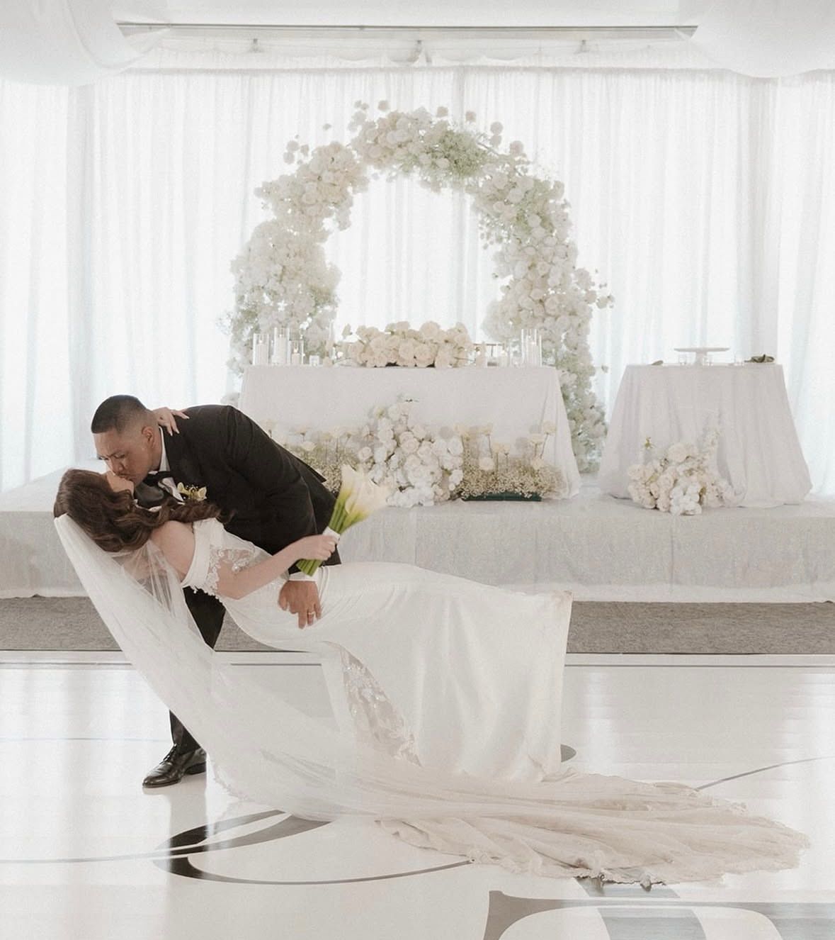 Newlyweds share a kiss during their wedding. Bride leans back, groom dips her. White floral arch and table.