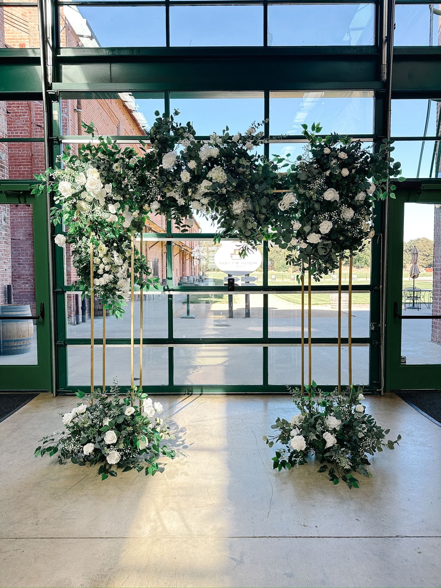 Wedding arch with white flowers and greenery, in front of a glass wall.