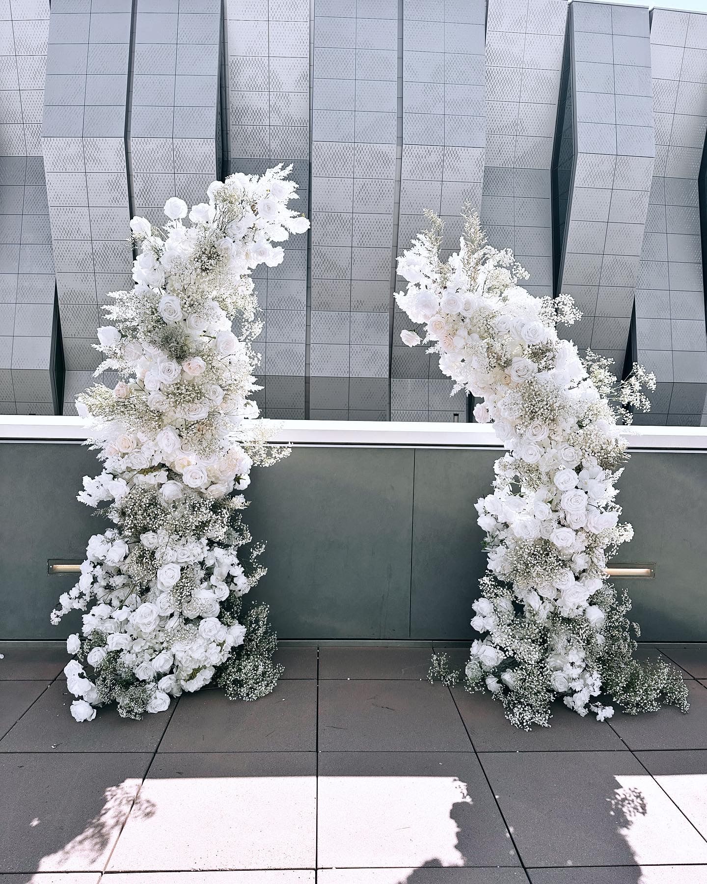 Two arches made of white flowers are in front of a brick wall