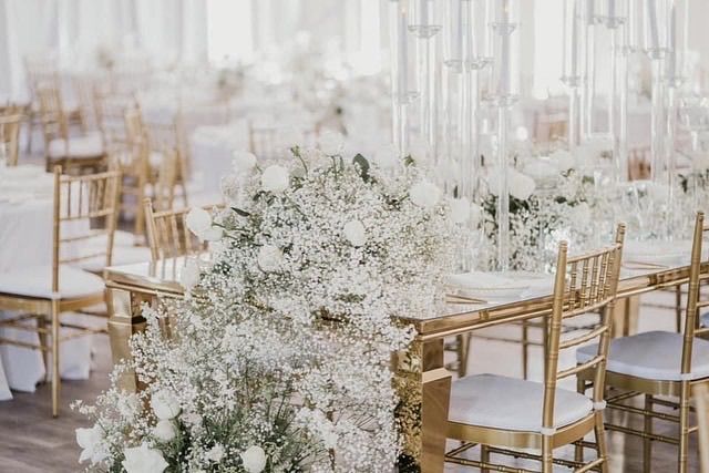 A long table with gold chairs and white flowers on it.